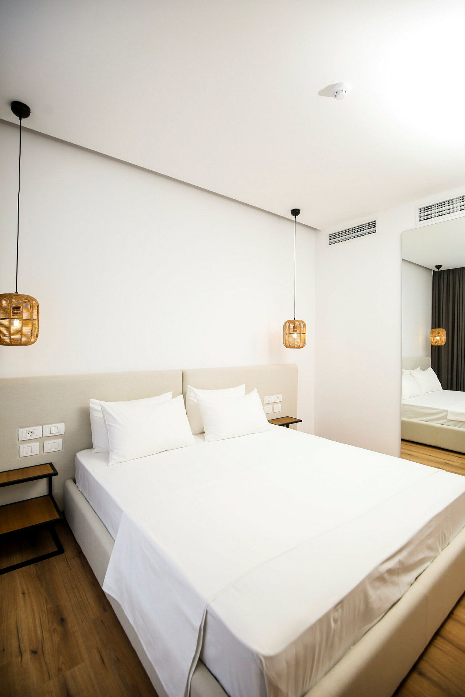 Et moderne hotellrom med en stor seng oppredet med hvite laken. Hengende lamper i rottingstil flankerer sengen. (AI-generert tekst)