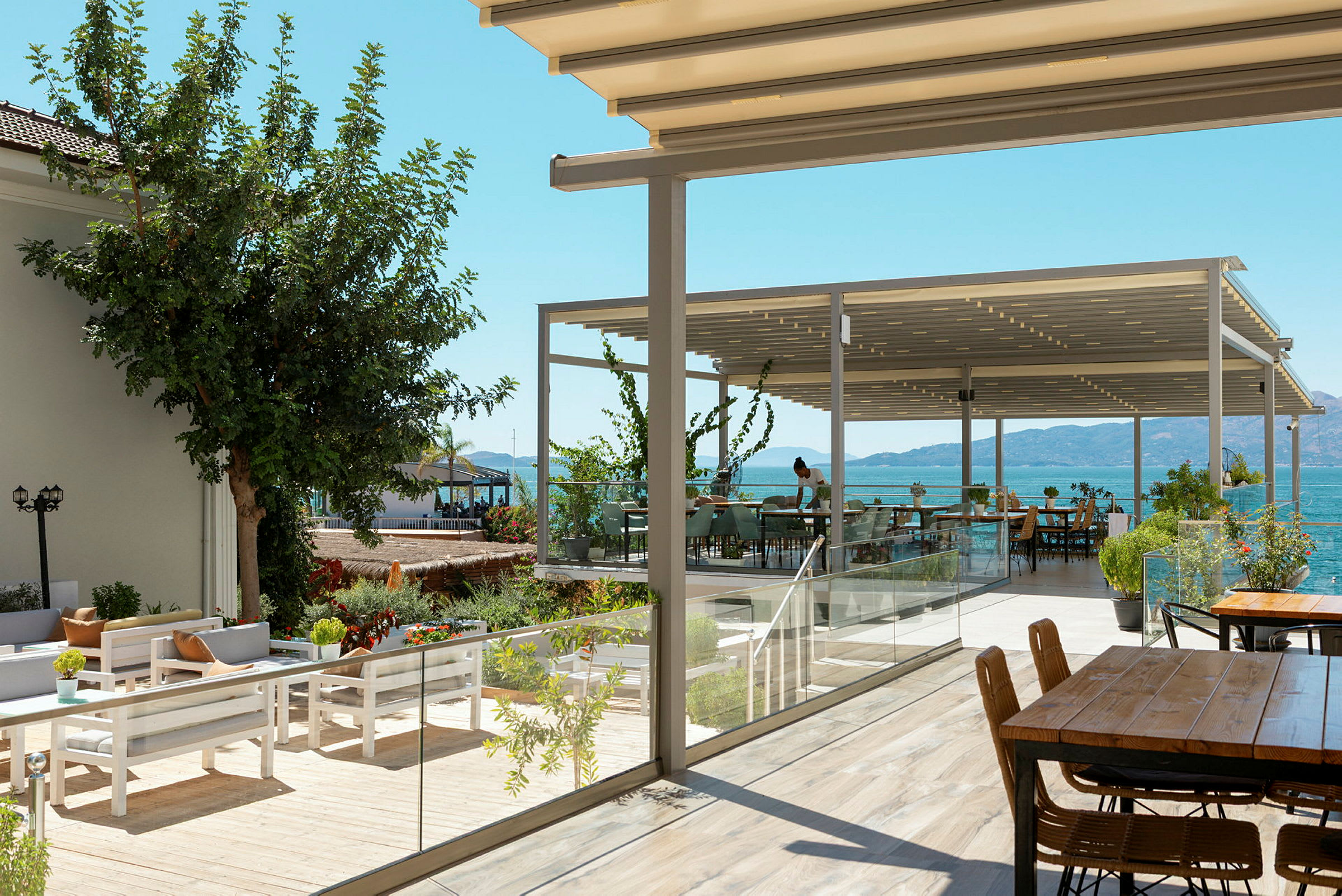 Bildet viser en utendørs servering med havutsikt og bord under en pergola. (AI-generert tekst)