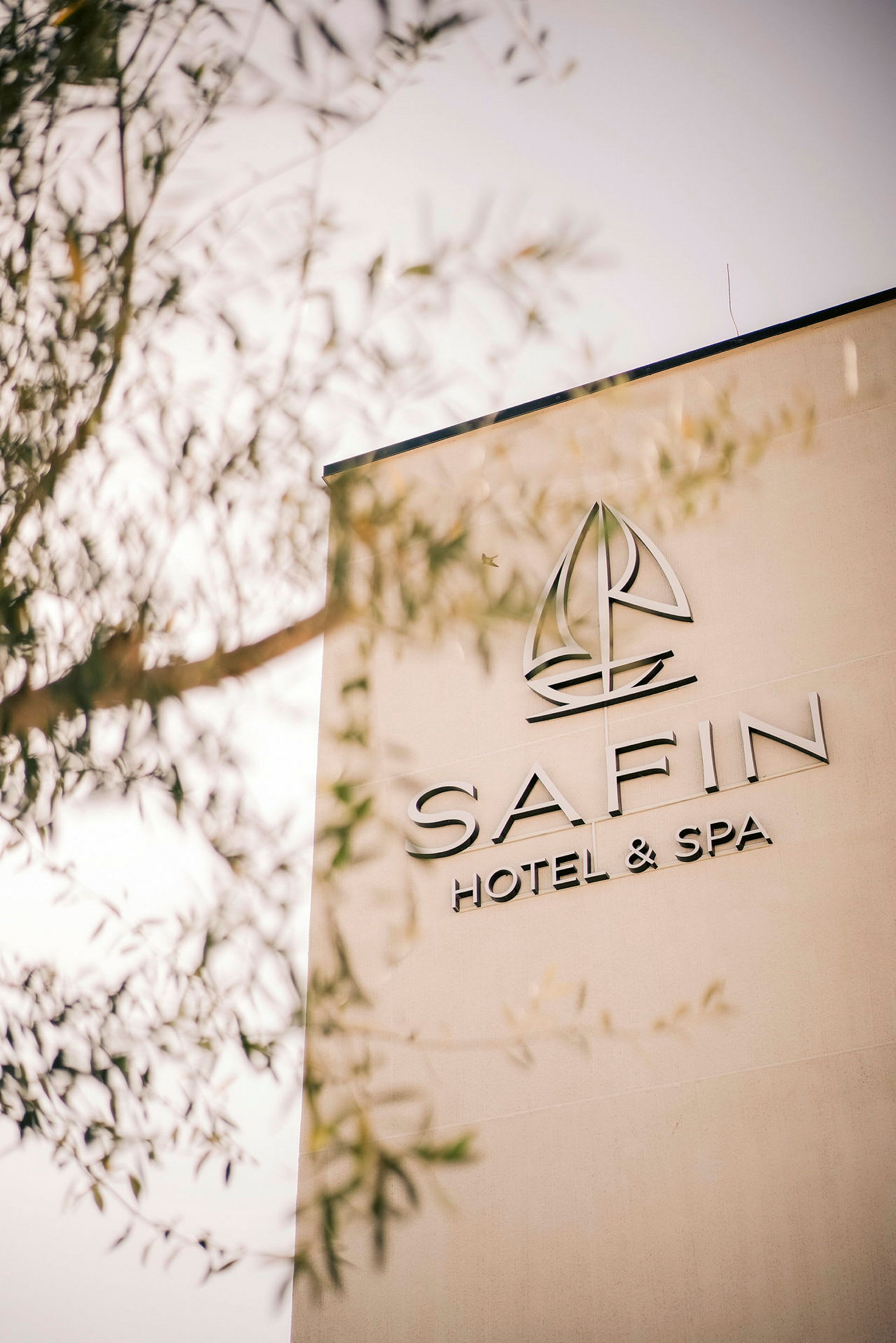 Bygningsvegg med teksten "SAFIN Hotel & Spa" og en logo som ligner en seilbåt. Deler av et tre skimtes i forgrunnen. (AI-generert tekst)