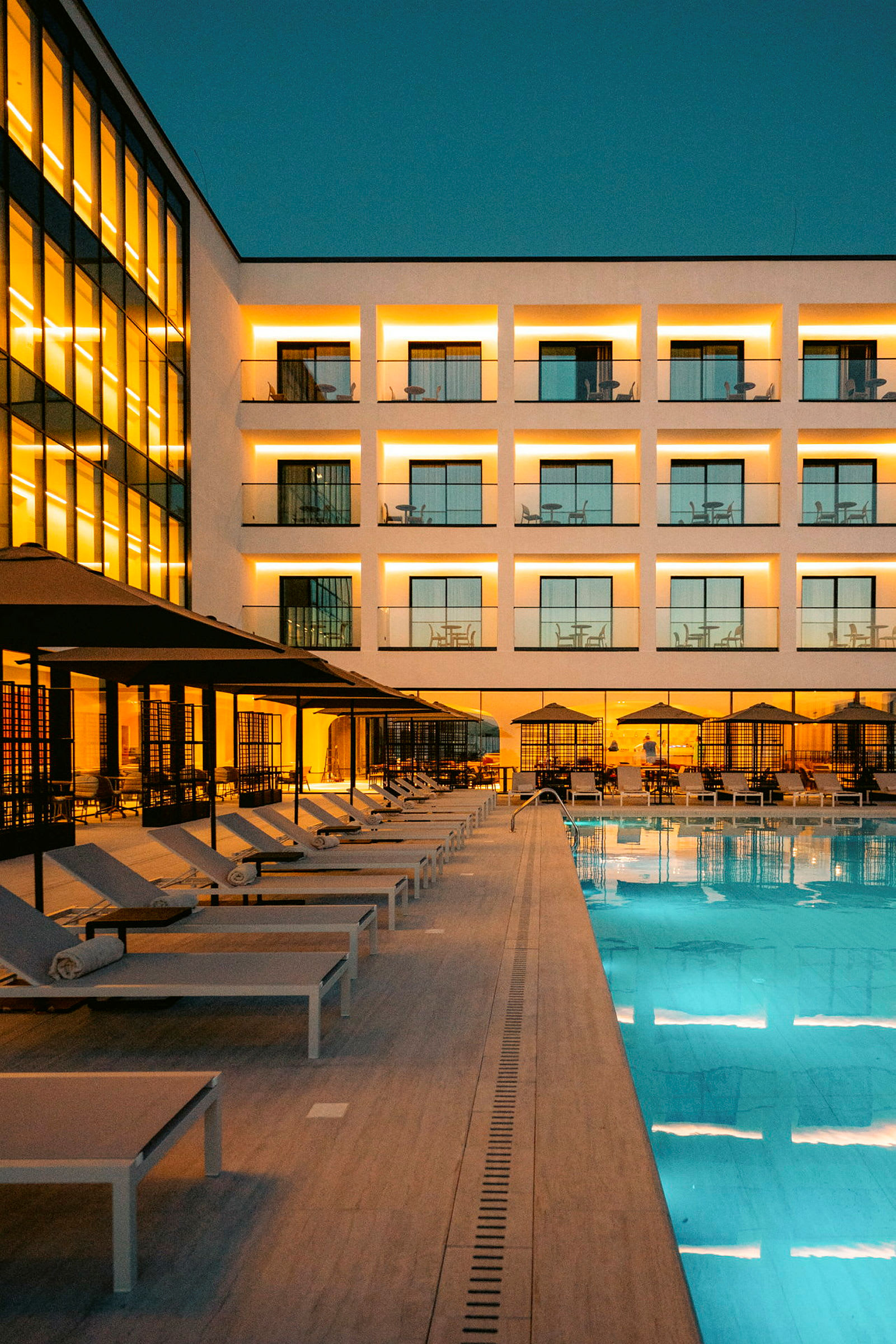 Bilde av en opplyst hotellfasade med balkonger, en rekke med solsenger under parasoller ved et svømmebasseng, under skumring. (AI-generert tekst)