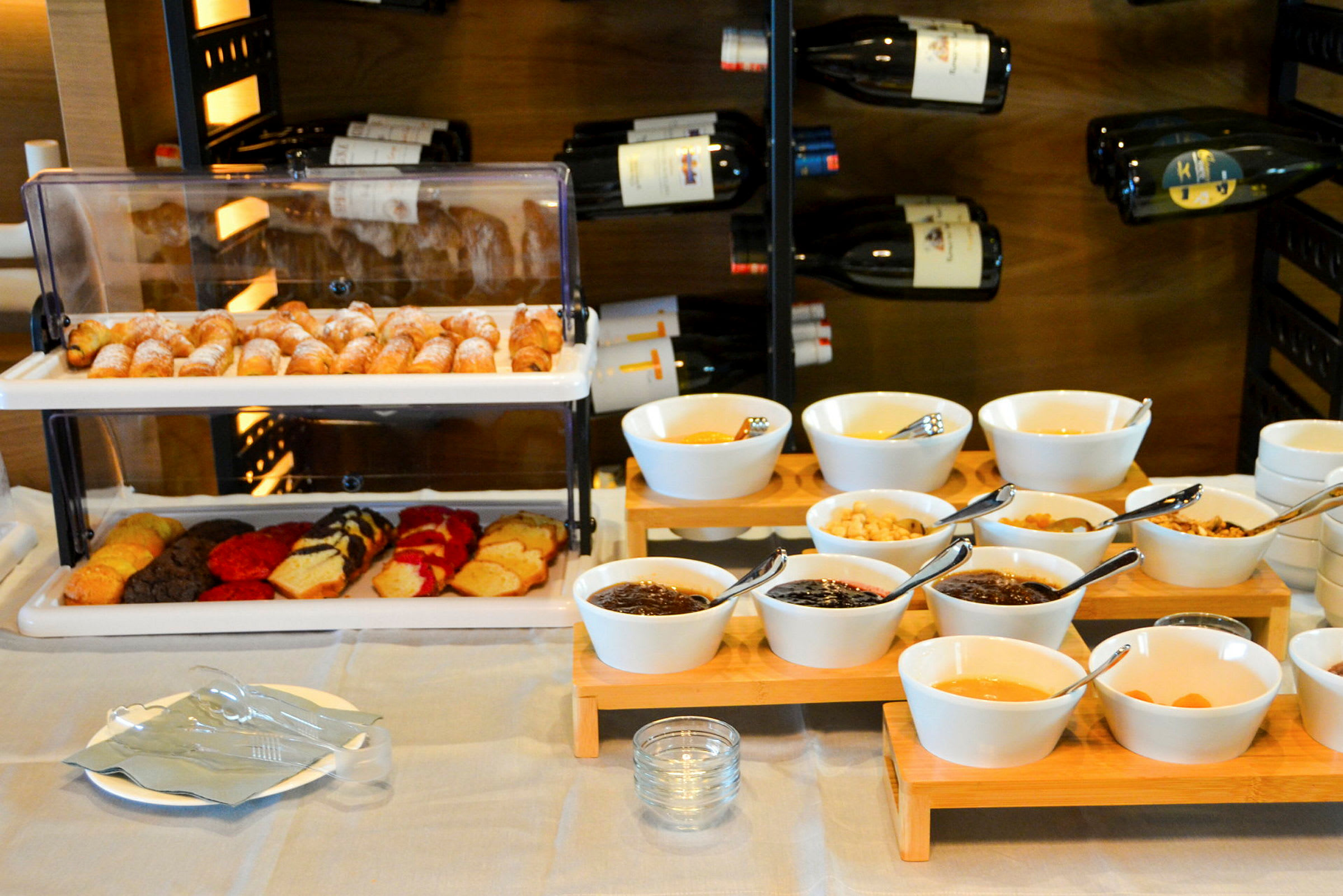 Et frokostbord med croissanter, kaker og skåler med forskjellige tilbehør foran en vinlagring. (AI-generert tekst)