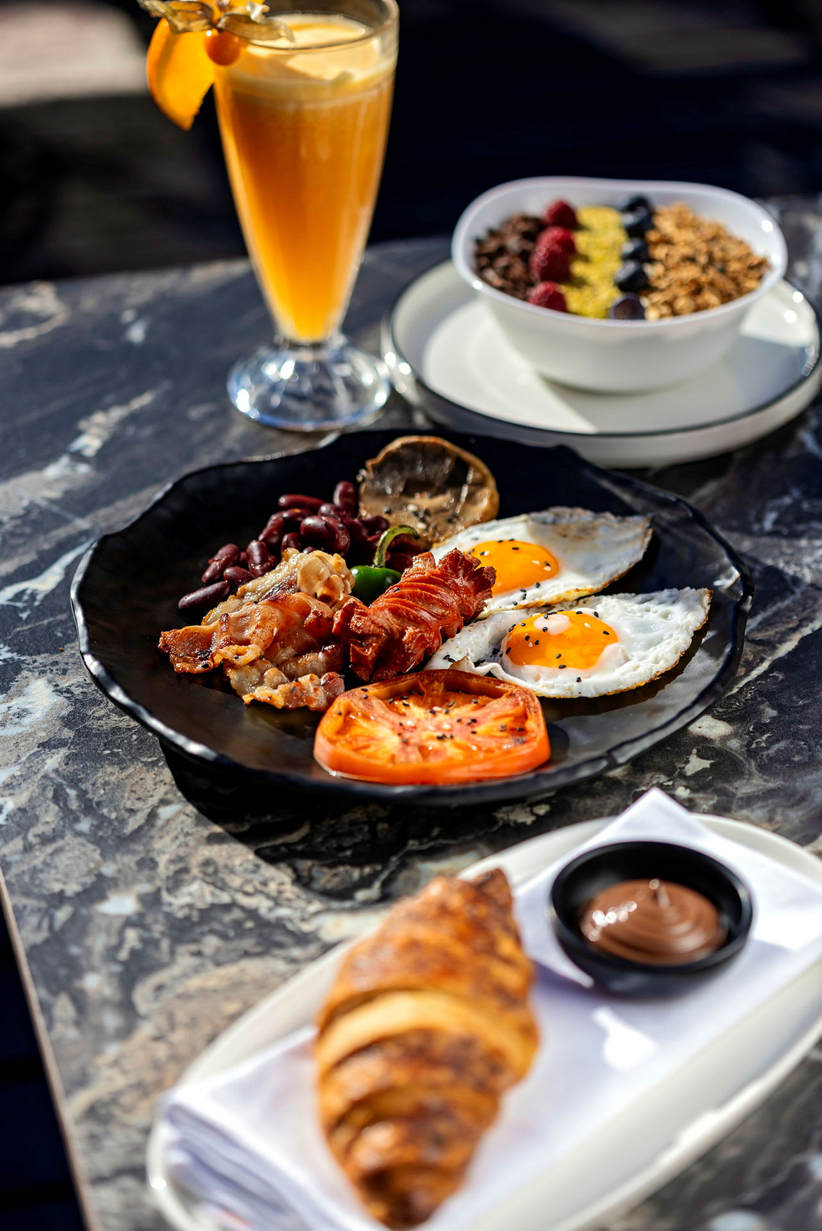 En tallerken med frokostmat bestående av bacon, egg, tomat og grønnsaker. En croissant med sjokoladekrem og en skål med frokostblanding samt et glass juice er også tilstede. (AI-generert tekst)