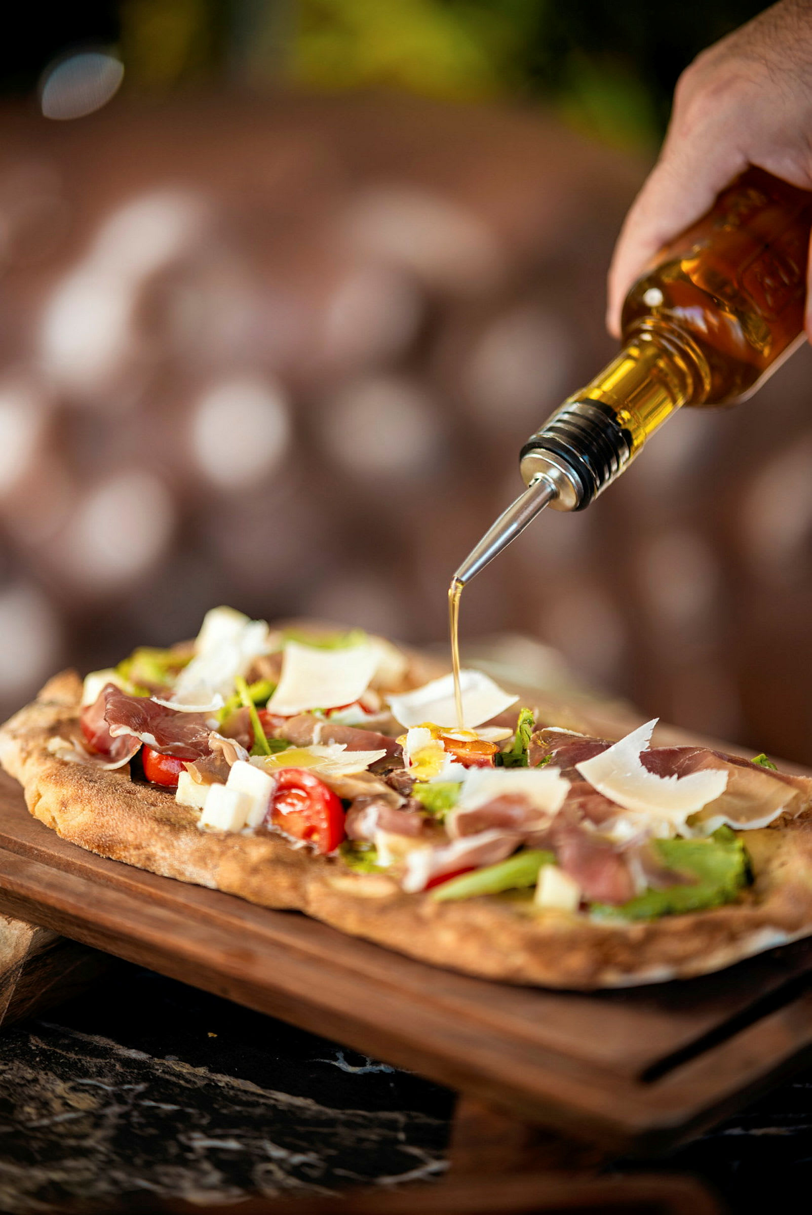 En hånd heller olivenolje fra en flaske over en pizza med skinke, tomater og parmesanost. (AI-generert tekst)