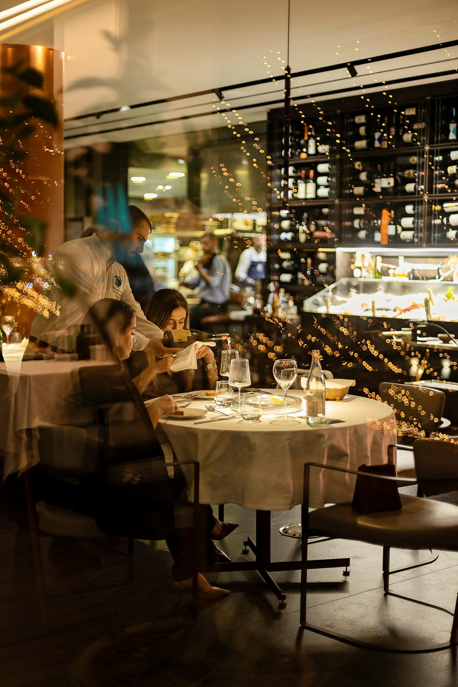 En restaurant med et bord dekket til måltid og en vinhylle i bakgrunnen. (AI-generert tekst)