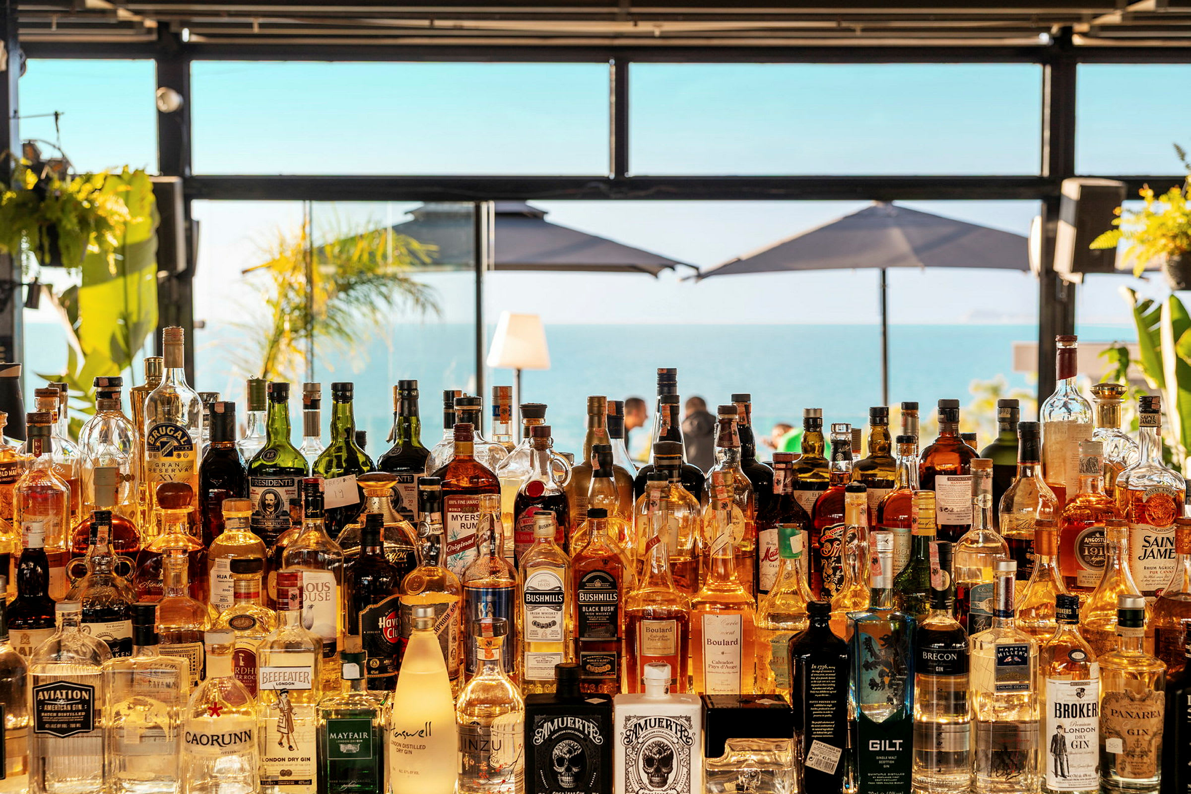 Utsikt over en strandbar med flasker på en hylle og havet i bakgrunnen. (AI-generert tekst)