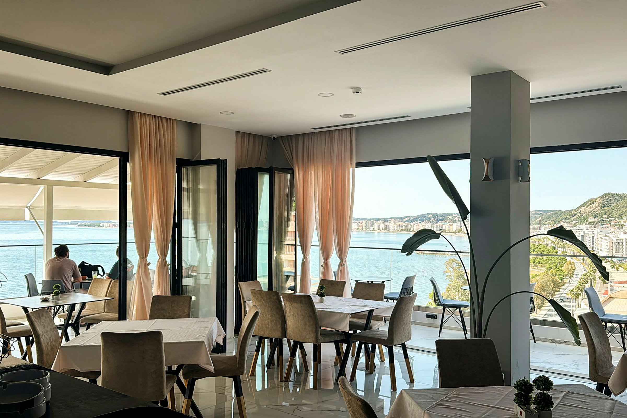 Et restaurantområde med store vinduer og utsikt over havet og en by. (AI-generert tekst)