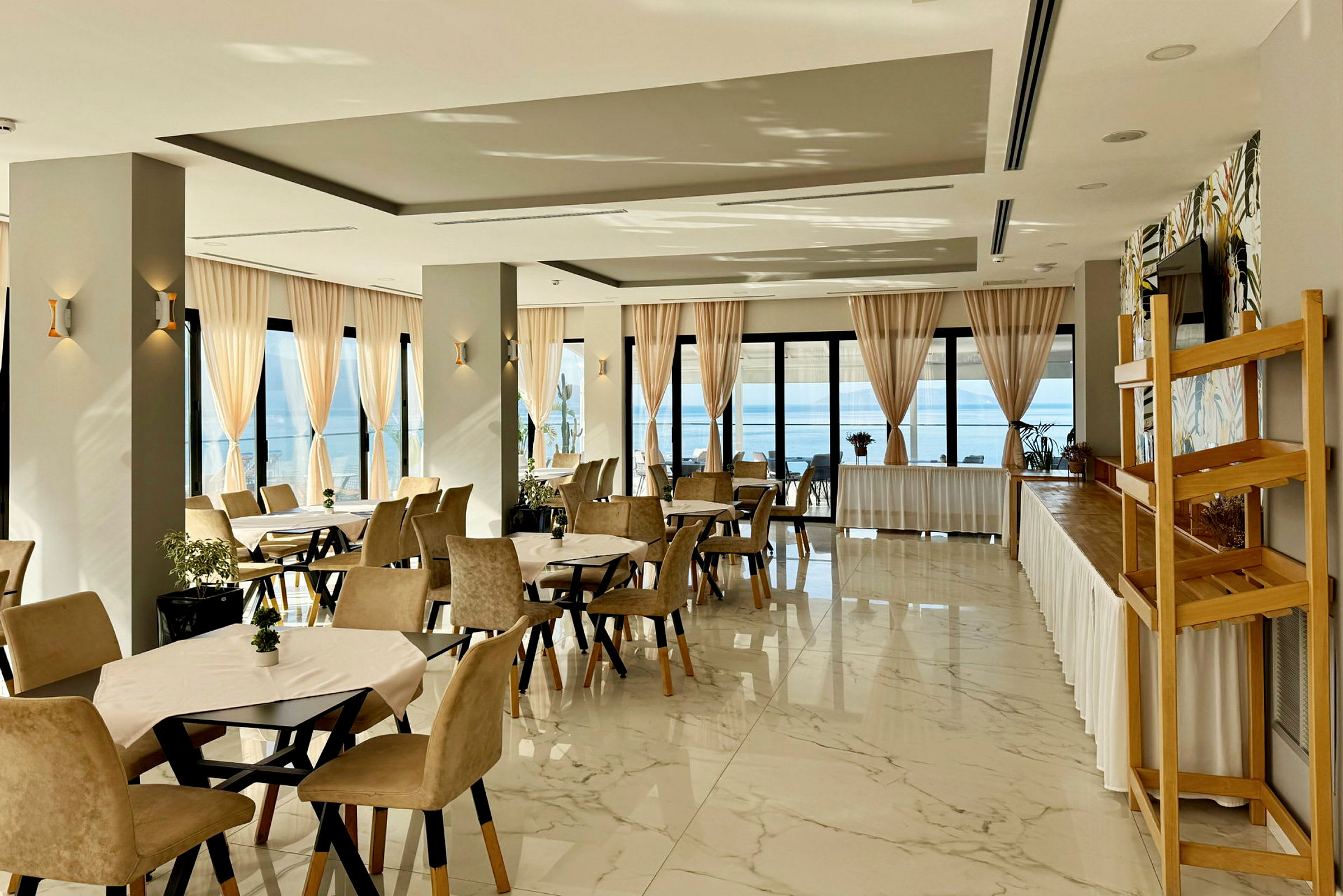 Restaurant med dekkede bord og beige stoler, store vinduer med lyse gardiner og havutsikt. (AI-generert tekst)