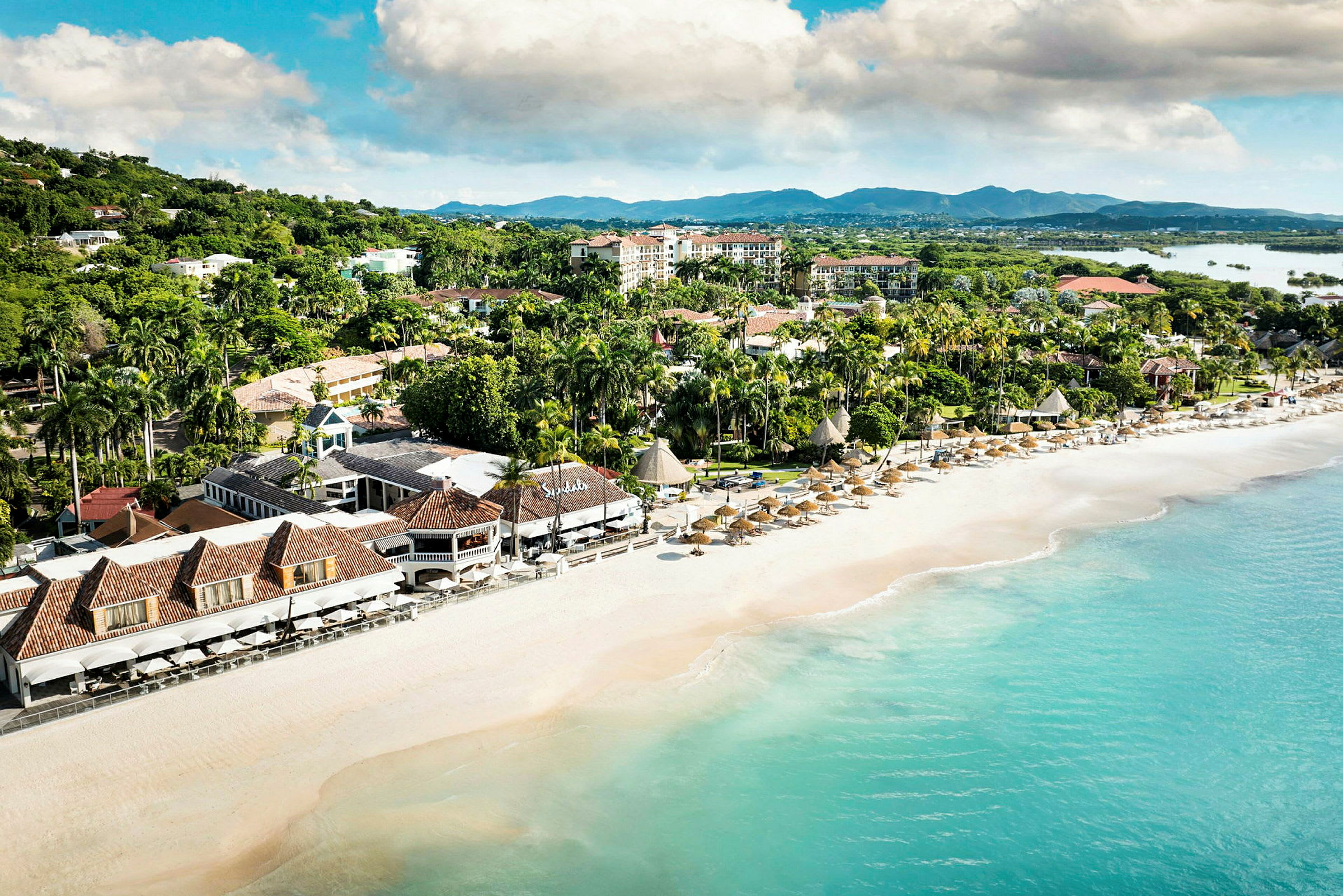 Flyfoto av en strand med parasoller og hotellbygninger omgitt av grøntområder. (AI-generert tekst)