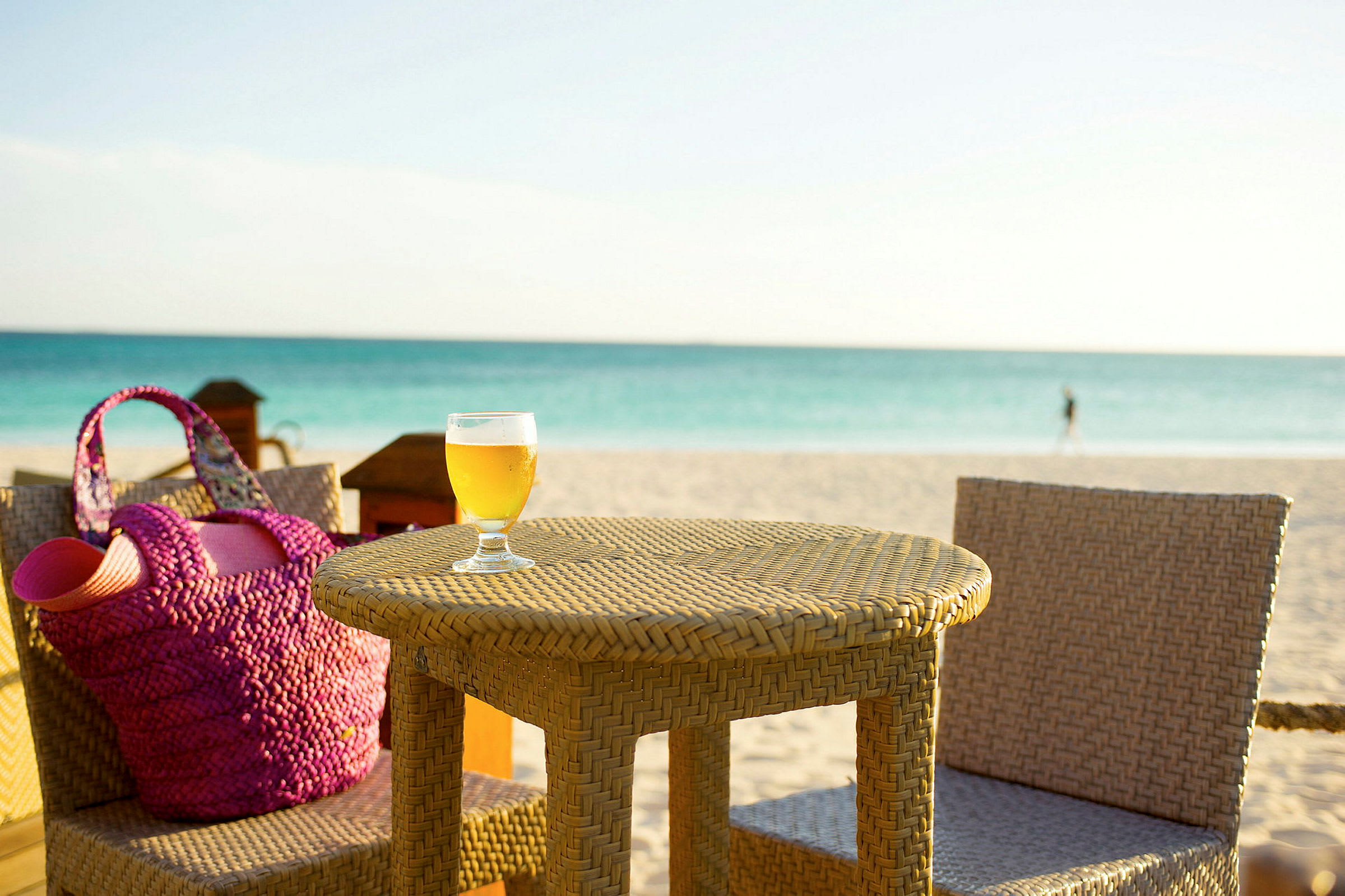 Et bord og stoler av flettet materiale står på en strand. På bordet står et glass med en drikk, og en rosa strandveske er synlig ved siden av. (AI-generert tekst)