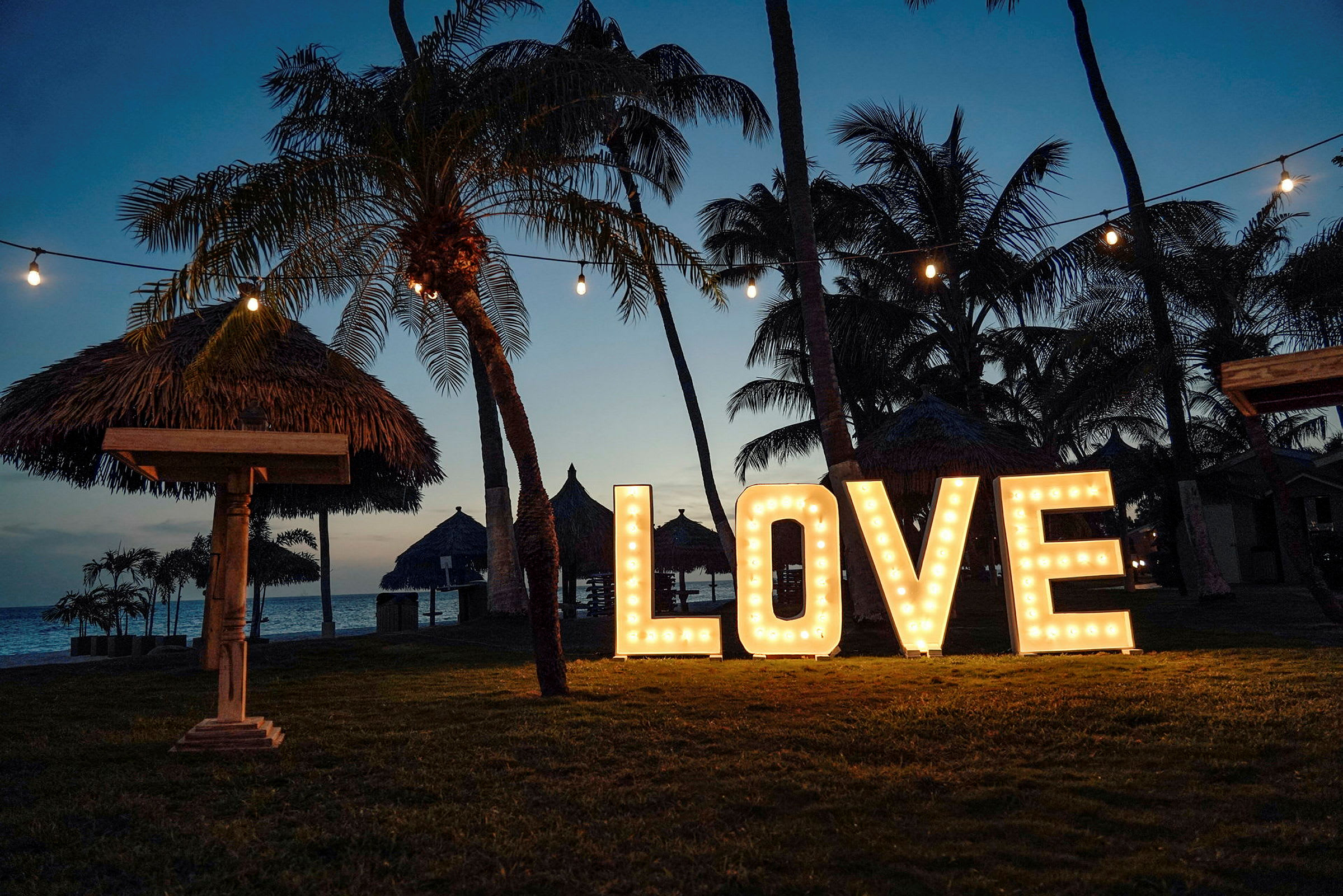 Store lysende bokstaver som danner ordet 'LOVE' står på en gressplen omgitt av palmer. Over naturscenen henger lyskjeder, og en strand kan ses i bakgrunnen i skumringen. (AI-generert tekst)
