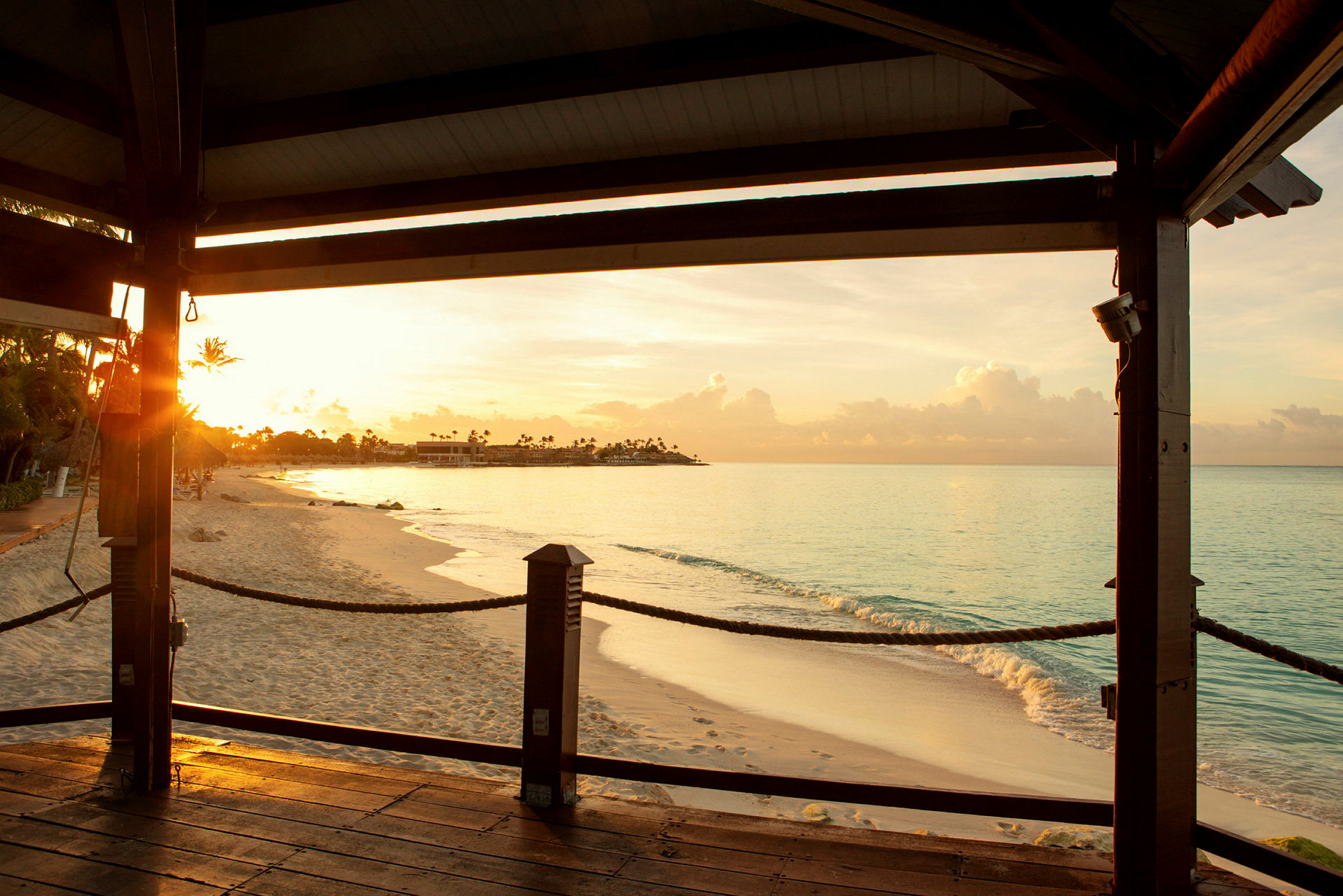 Bildet viser en strand ved solnedgang, sett fra en trebrygge under et tak. (AI-generert tekst)