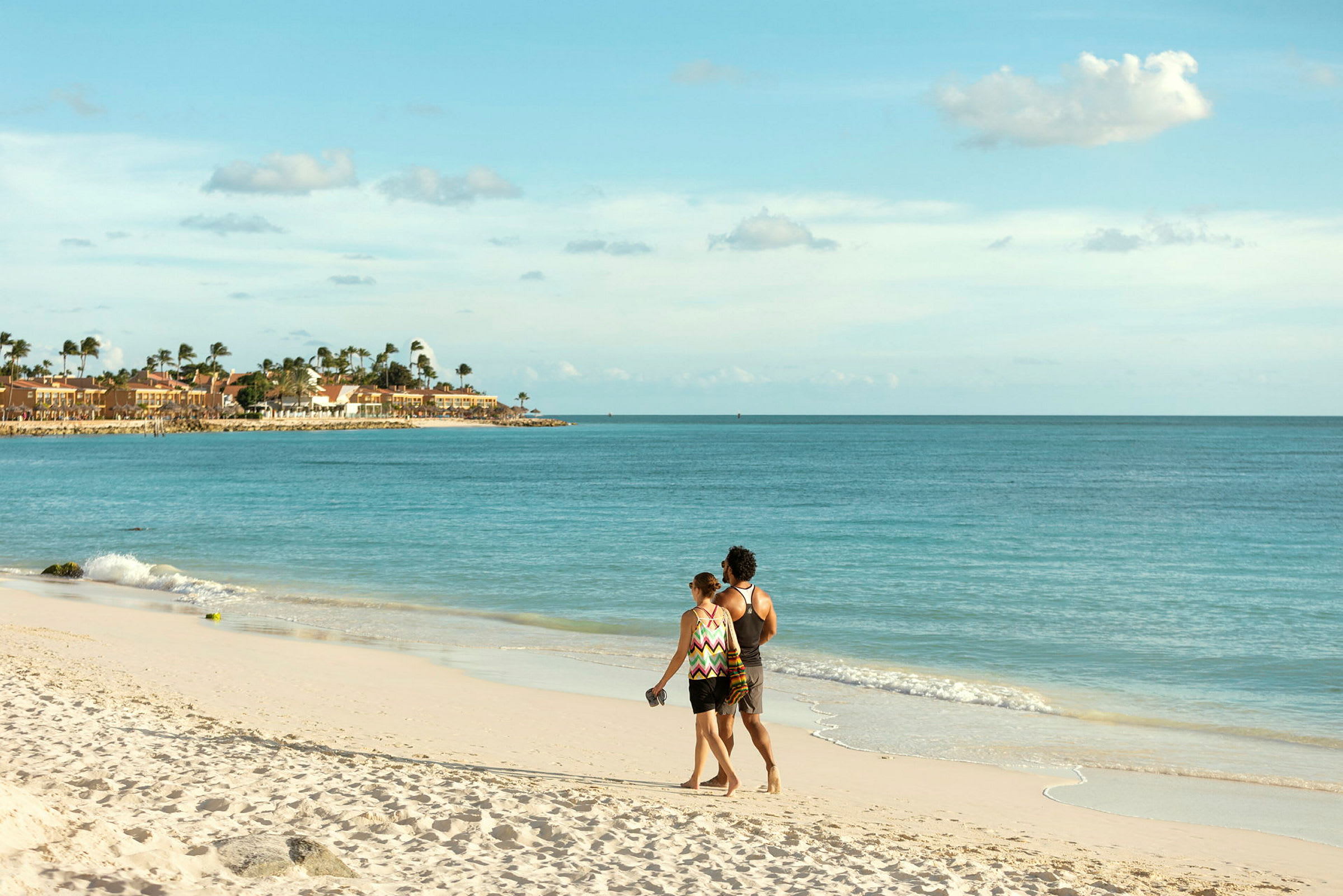 Et par går langs en sandstrand med utsikt over havet og bygninger langs kysten. (AI-generert tekst)