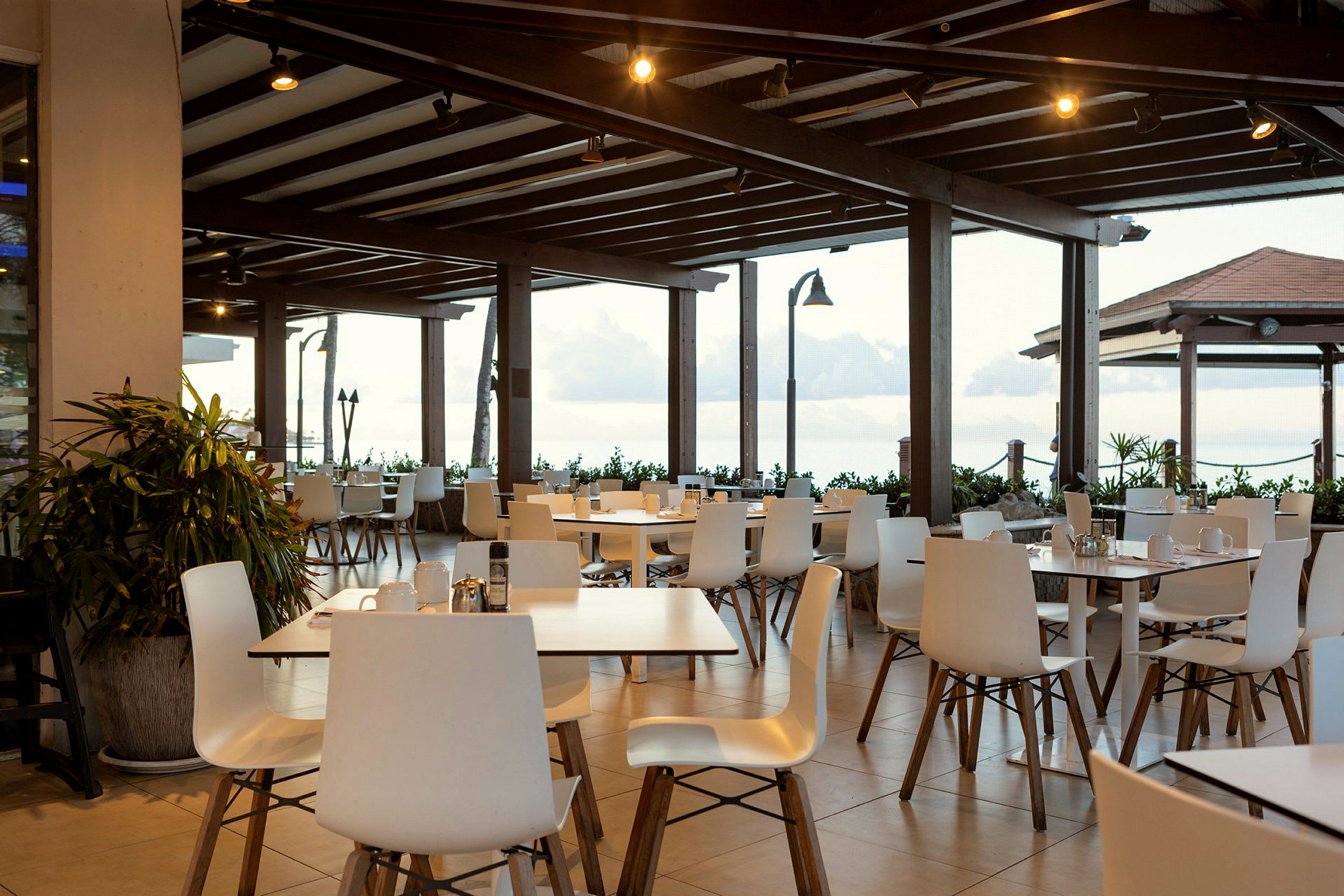 En restaurantterrasse med hvite stoler og bord under et tretak. Utsikt over havet bak. (AI-generert tekst)