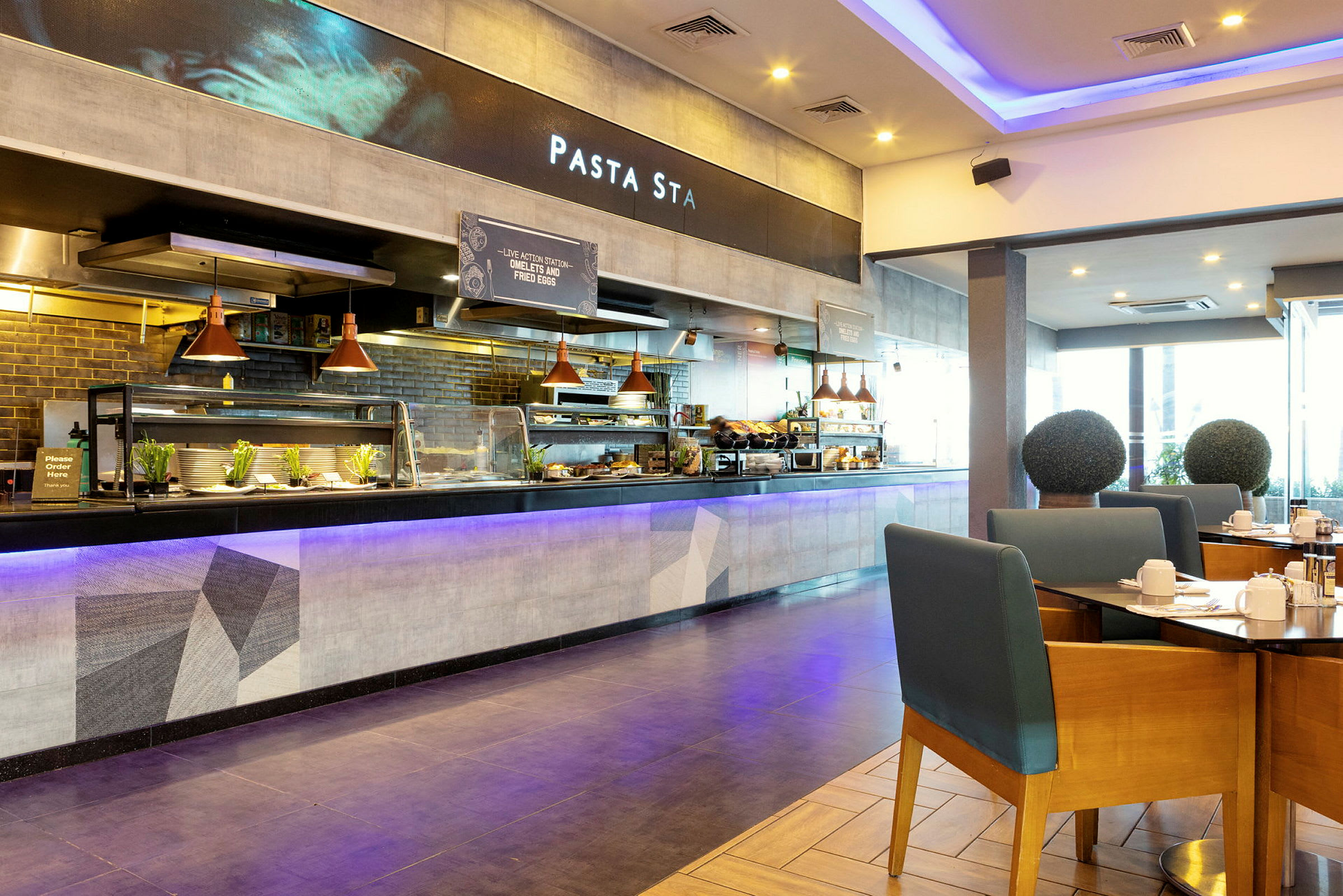 En restaurant med en åpen serveringsdisk og sitteplasser. Bord med stoler er synlige i forgrunnen, og bak serveringsdisken vises et skilt med teksten 'Pasta Sta'. (AI-generert tekst)