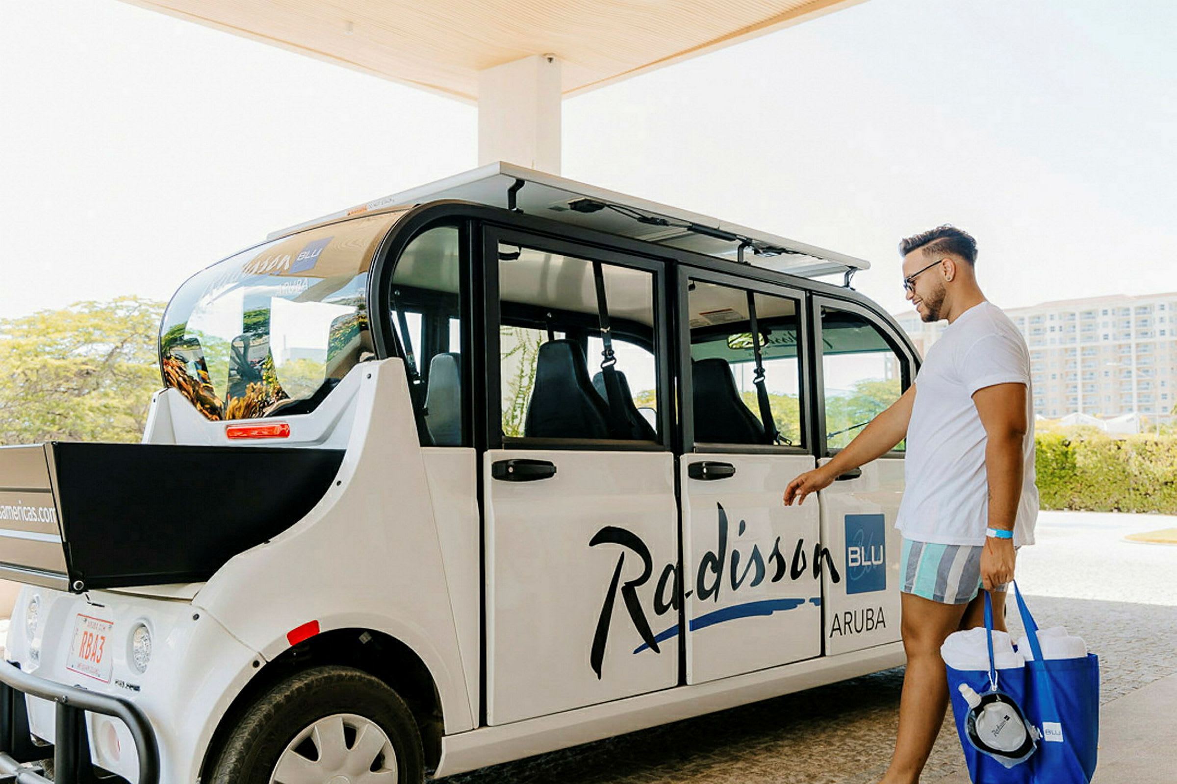 En turist står ved siden av en hvit transportbil med teksten 'Radisson Blu Aruba'. (AI-generert tekst)