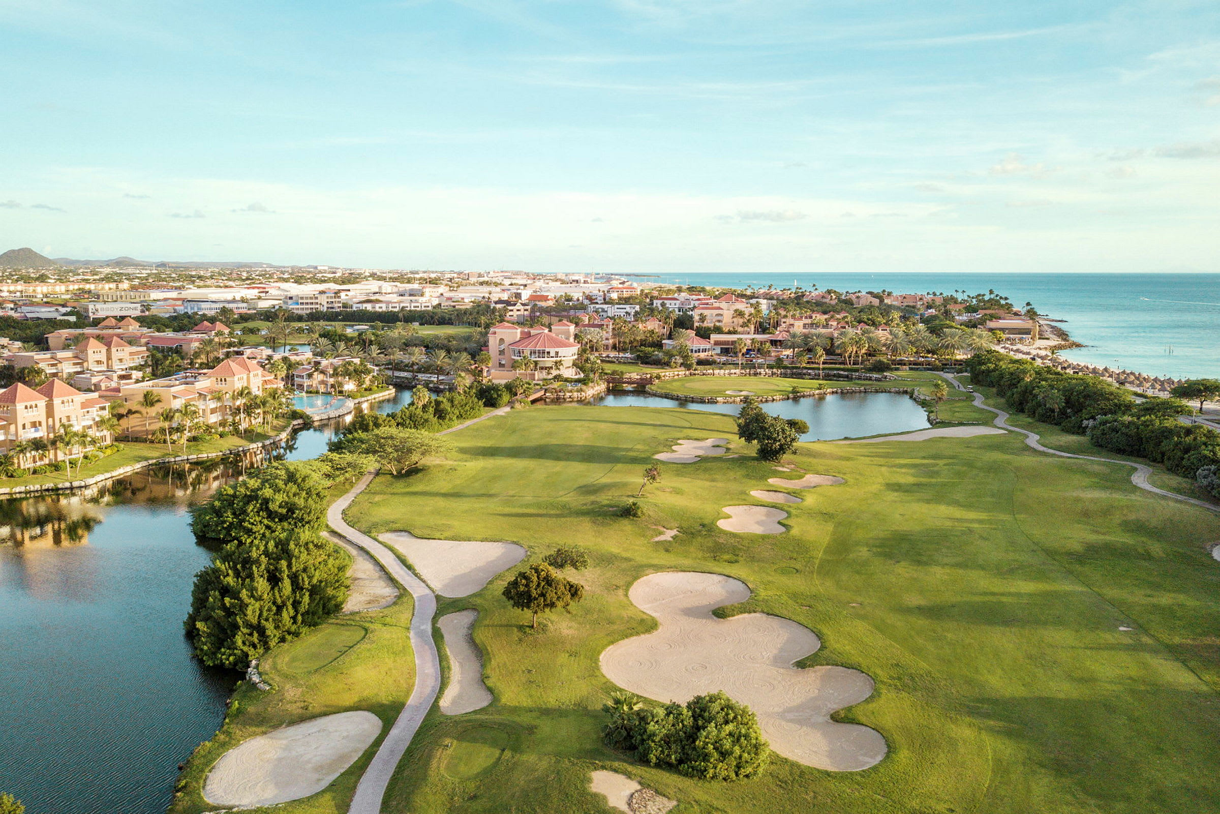 Golfbane med sandbunkere og grønne plenområder nær vannløp og strand. Bygninger i bakgrunnen. (AI-generert tekst)