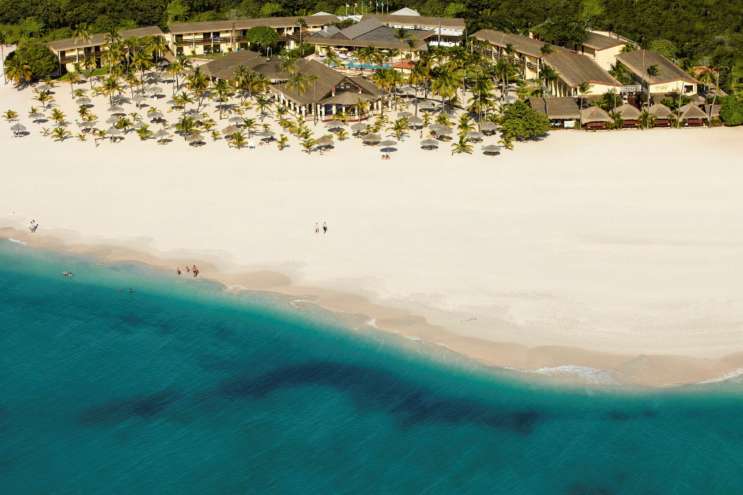 Flyfoto av en hvit sandstrand med blått hav foran et hotellområde omgitt av palmer. (AI-generert tekst)