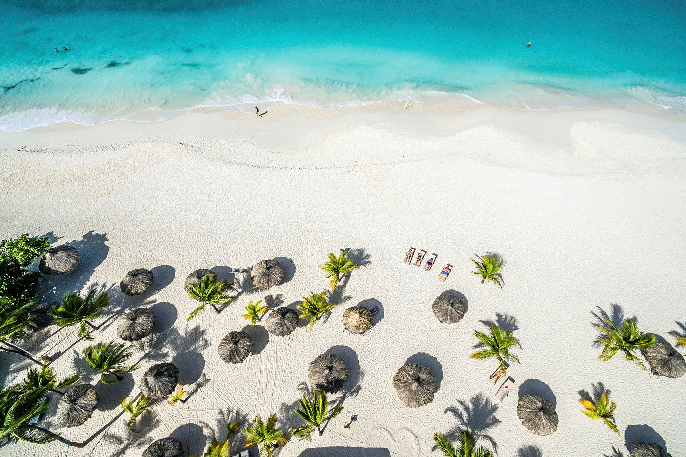 Luftfoto av en hvit sandstrand med turkis vann. Stranden er dekorert med parasoller og omgitt av palmer. (AI-generert tekst)