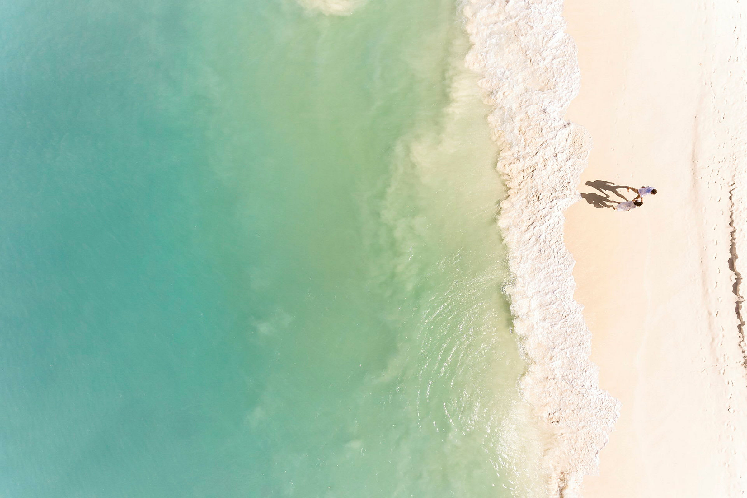 Et luftbilde av en strand med lys sand der bølger fra et turkis hav møter stranden. Et par personer går på stranden. (AI-generert tekst)