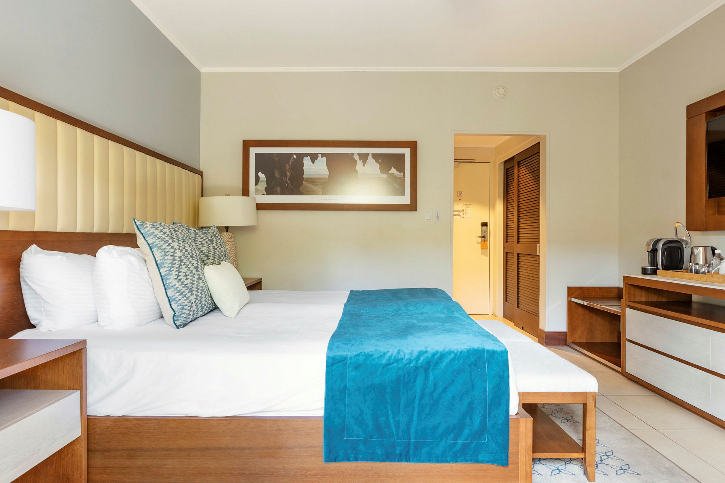 Et hotellrom med en dobbeltseng med hvitt sengetøy og et turkis sengeteppesett. Et skrivebord med kaffemaskin er til høyre. (AI-generert tekst)