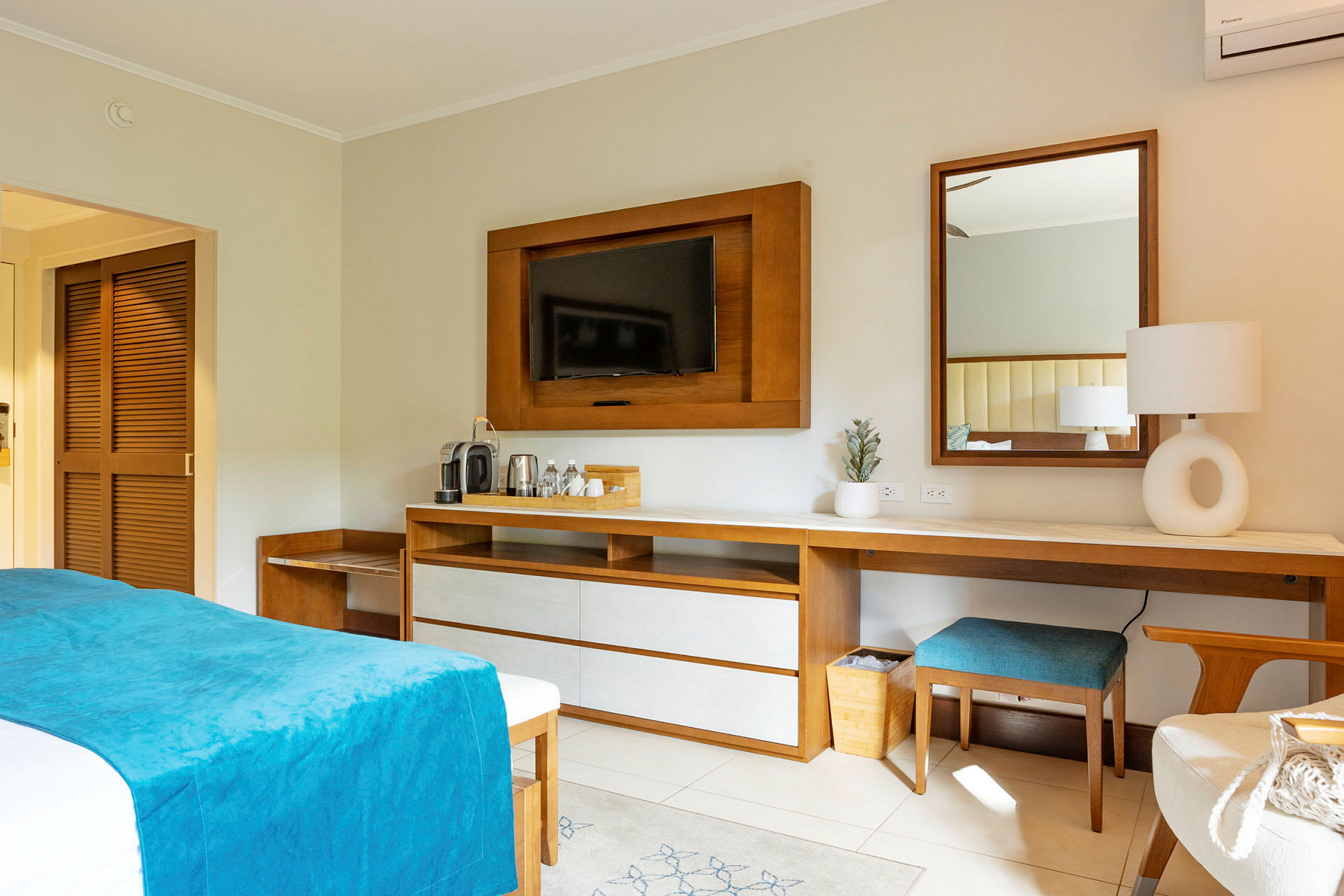 Et hotellrom med en seng med blått sengeteppe, tremøbler, en TV på veggen og et skrivebord med speil og lampe. (AI-generert tekst)
