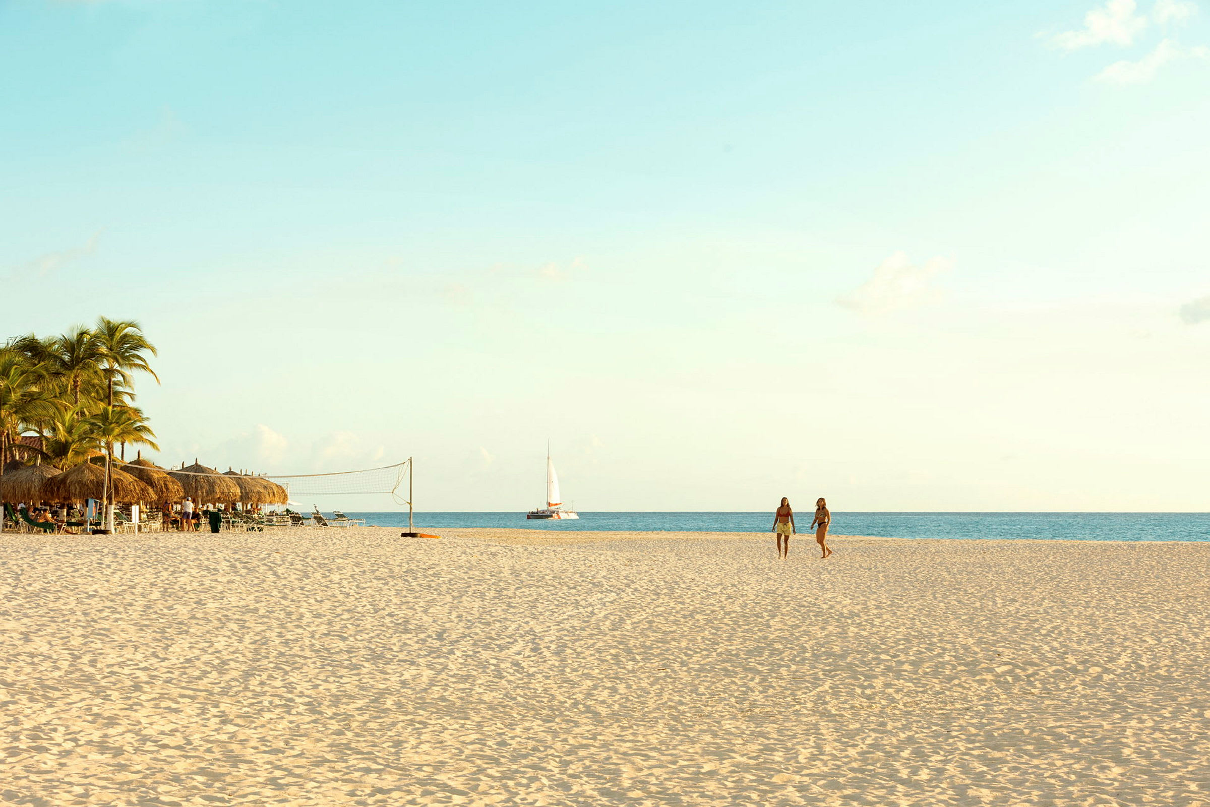 En sandstrand med palmer og parasoller til venstre, to personer som går på stranden og en seilbåt på havet. (AI-generert tekst)