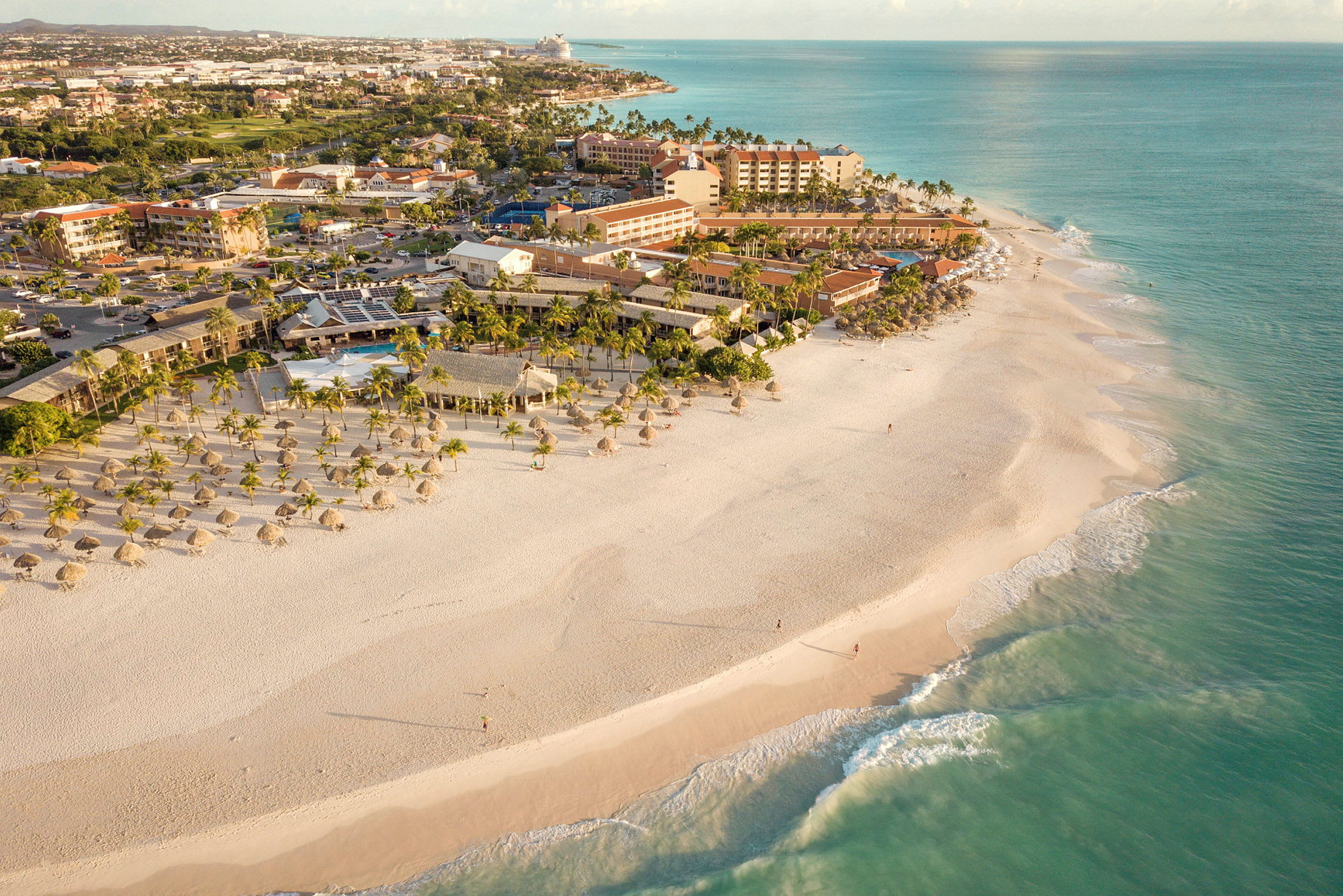 En strand med hvit sand med parasoller og palmer, og bak stranden vises bygninger og vegetasjon. (AI-generert tekst)