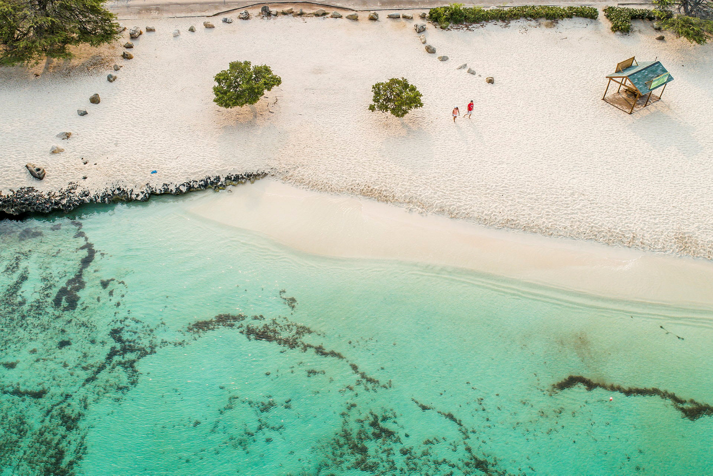 En strand med hvit sand, grønne busker og klart turkis vann vises på bildet. To personer går på stranden ved siden av en liten trehytte. (AI-generert tekst)