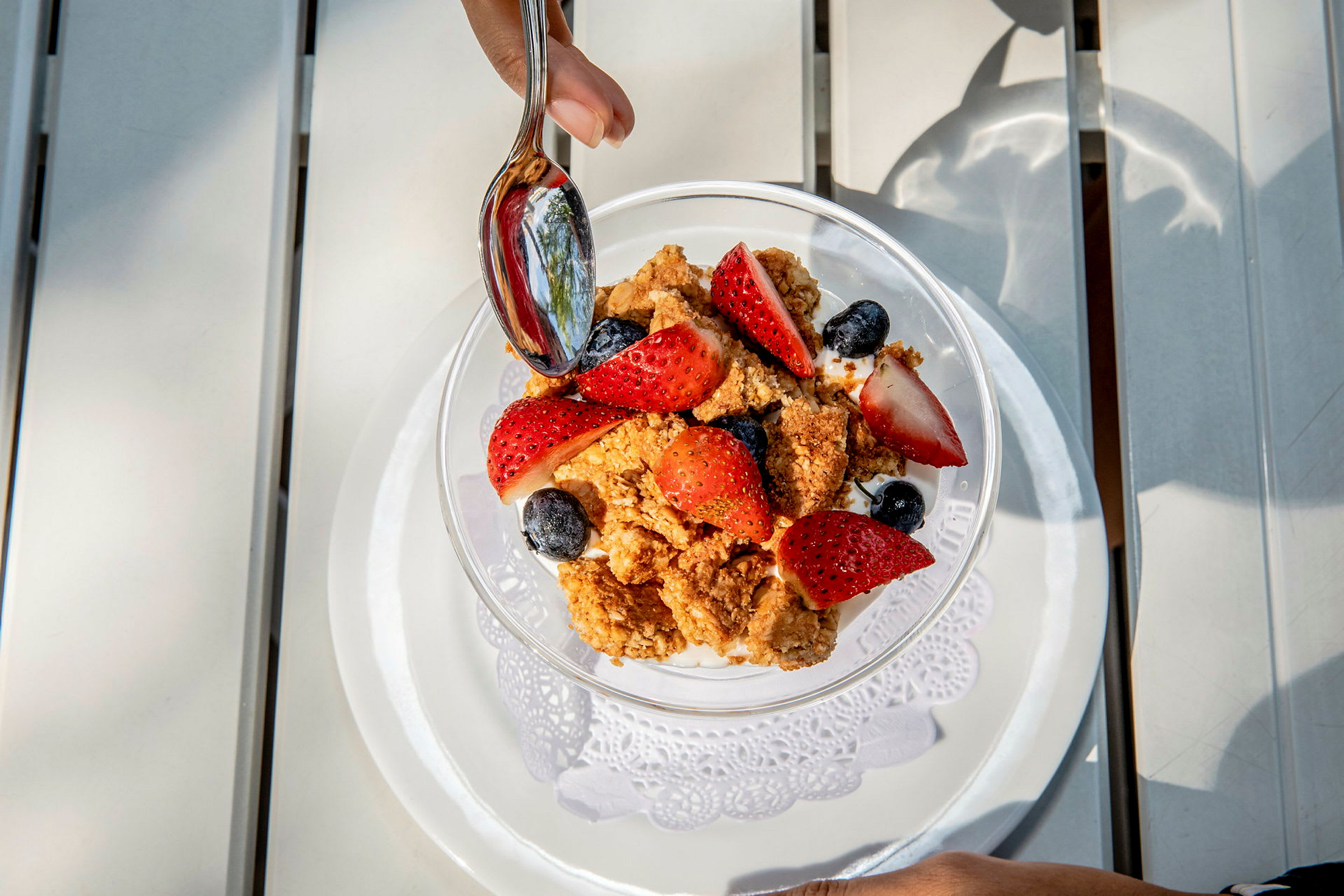 En bolle med yoghurt, ferske jordbær, blåbær og sprø granola. En hånd holder en skje over maten. (AI-generert tekst)