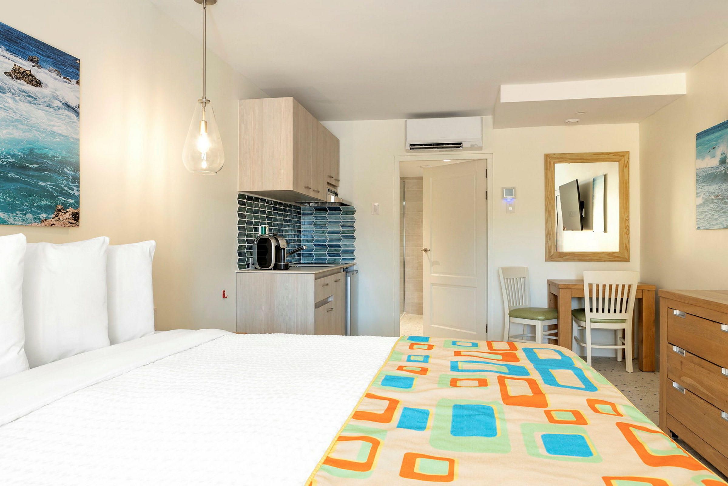 Et hotellrom med en seng i forgrunnen, et kjøkken og et bord med stoler i bakgrunnen. Veggene er dekorert med malerier. (AI-generert tekst)