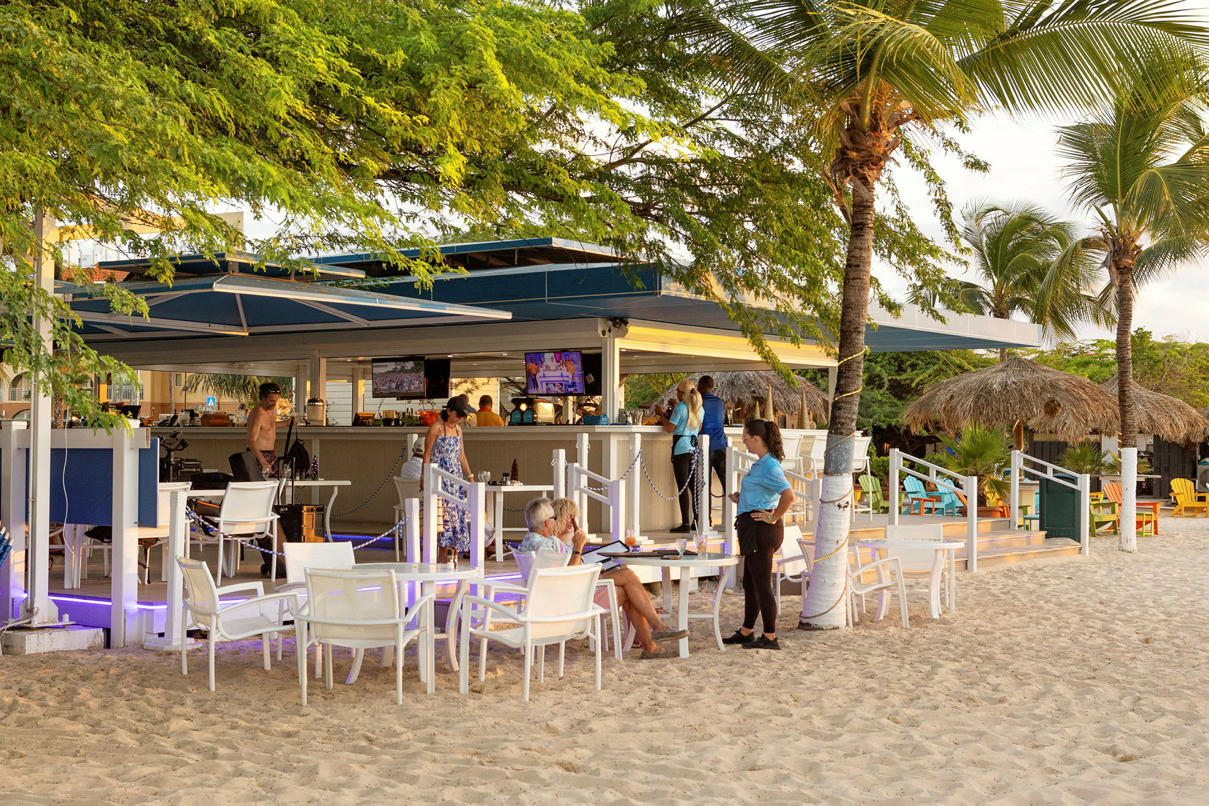 En utendørs strandbar med bord og stoler på sanden, omringet av grøntområder og trær. (AI-generert tekst)