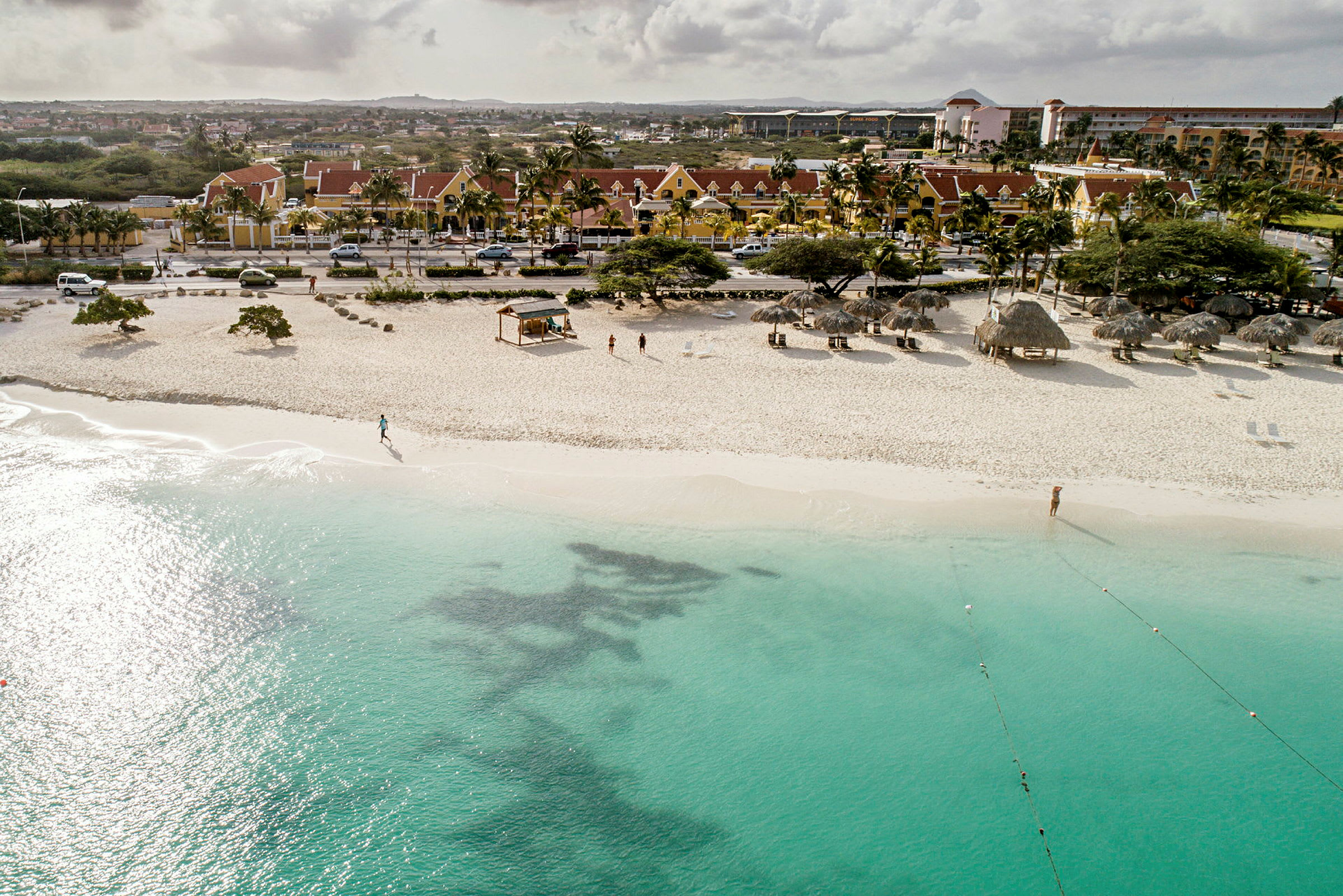 Sandstrand med turkis vann, parasoller og noen personer på stranden, samt bygninger i bakgrunnen. (AI-generert tekst)