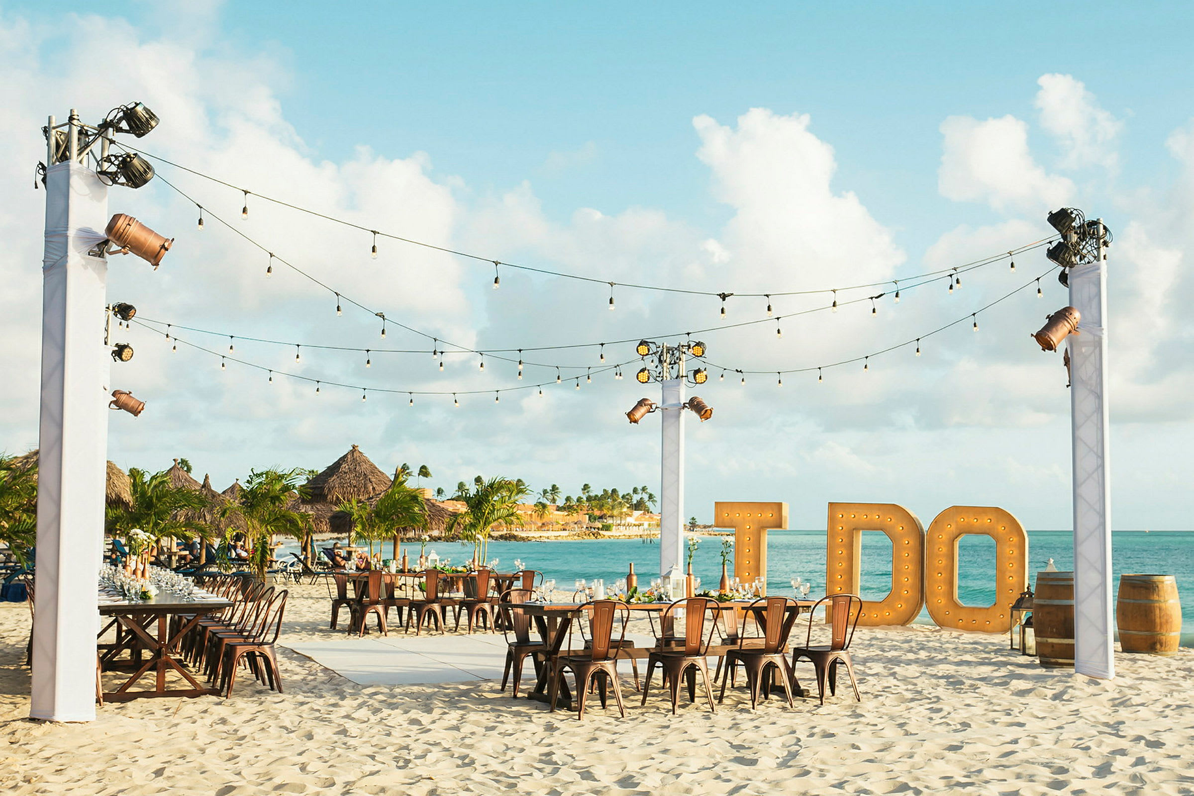 Et strandbryllup med bord og stoler arrangert på sanden, dekorasjoner med lyslenker og store bokstaver som danner teksten 'I DO' i bakgrunnen. (AI-generert tekst)