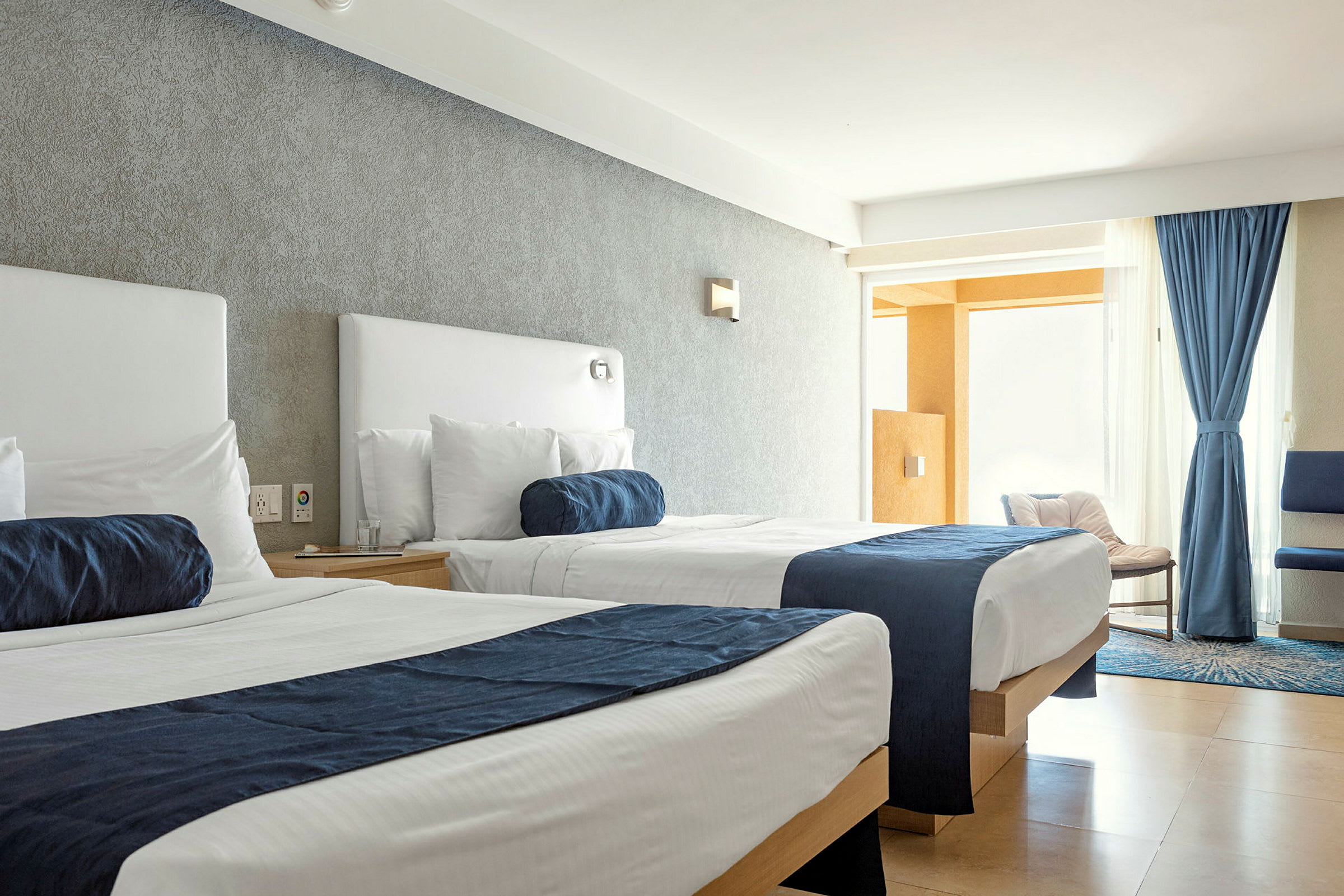 Et hotellrom med to senger med hvite laken og blå sengetepper. Vinduet er dekket med blå gardiner og lys strømmer inn i rommet. (AI-generert tekst)