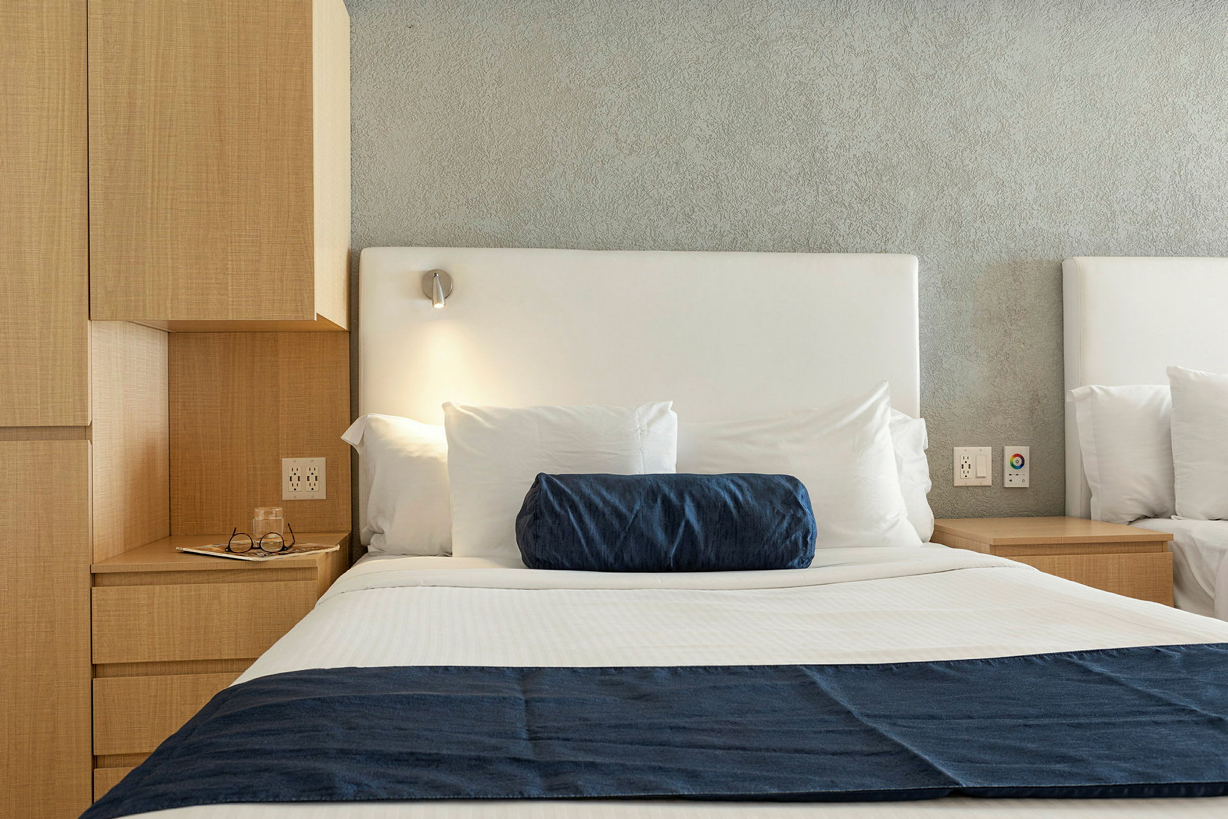 Bilde av en hotellseng med hvitt sengetøy, en blå pyntepute og et blått sengeteppe samt en sengeplampe over. (AI-generert tekst)