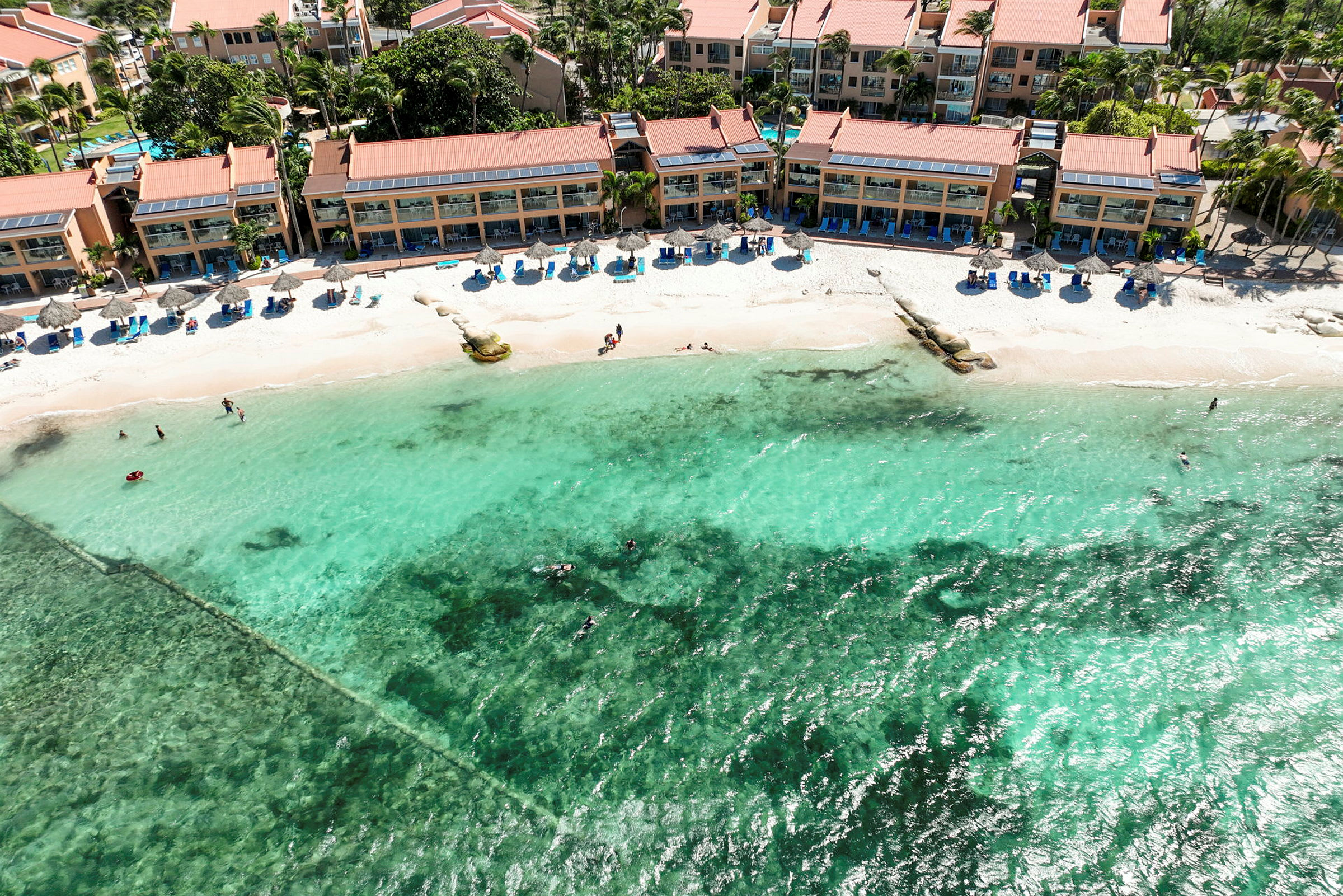 Luftfoto av en strand med turkist vann, omgitt av hotellbygninger og solsenger. (AI-generert tekst)