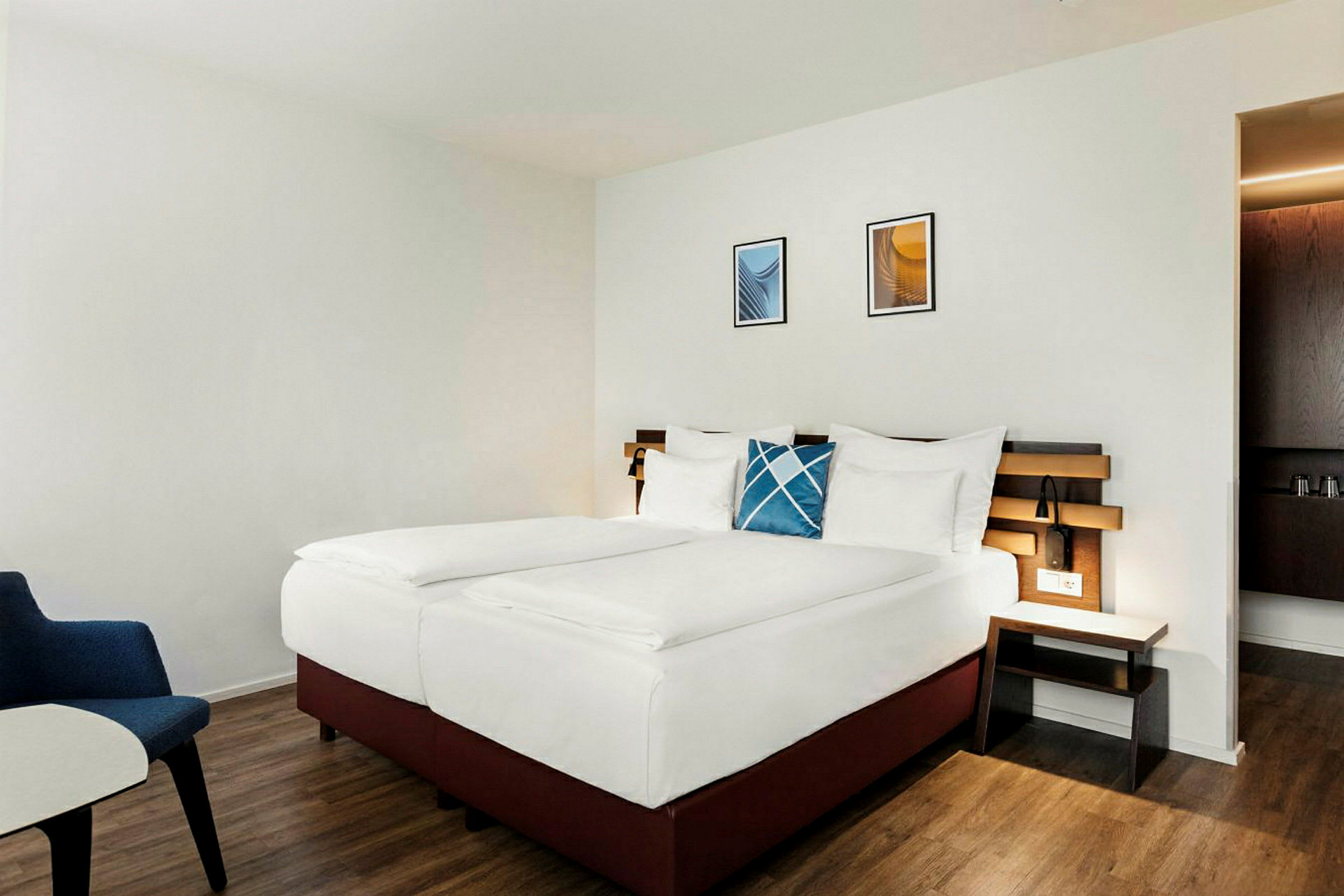 Hotellrom med en dobbeltseng, hvite sengetrekk og en blå dekorativ pute. En trepanel fungerer som sengegavl, og rommet har tregulv. (AI-generert tekst)