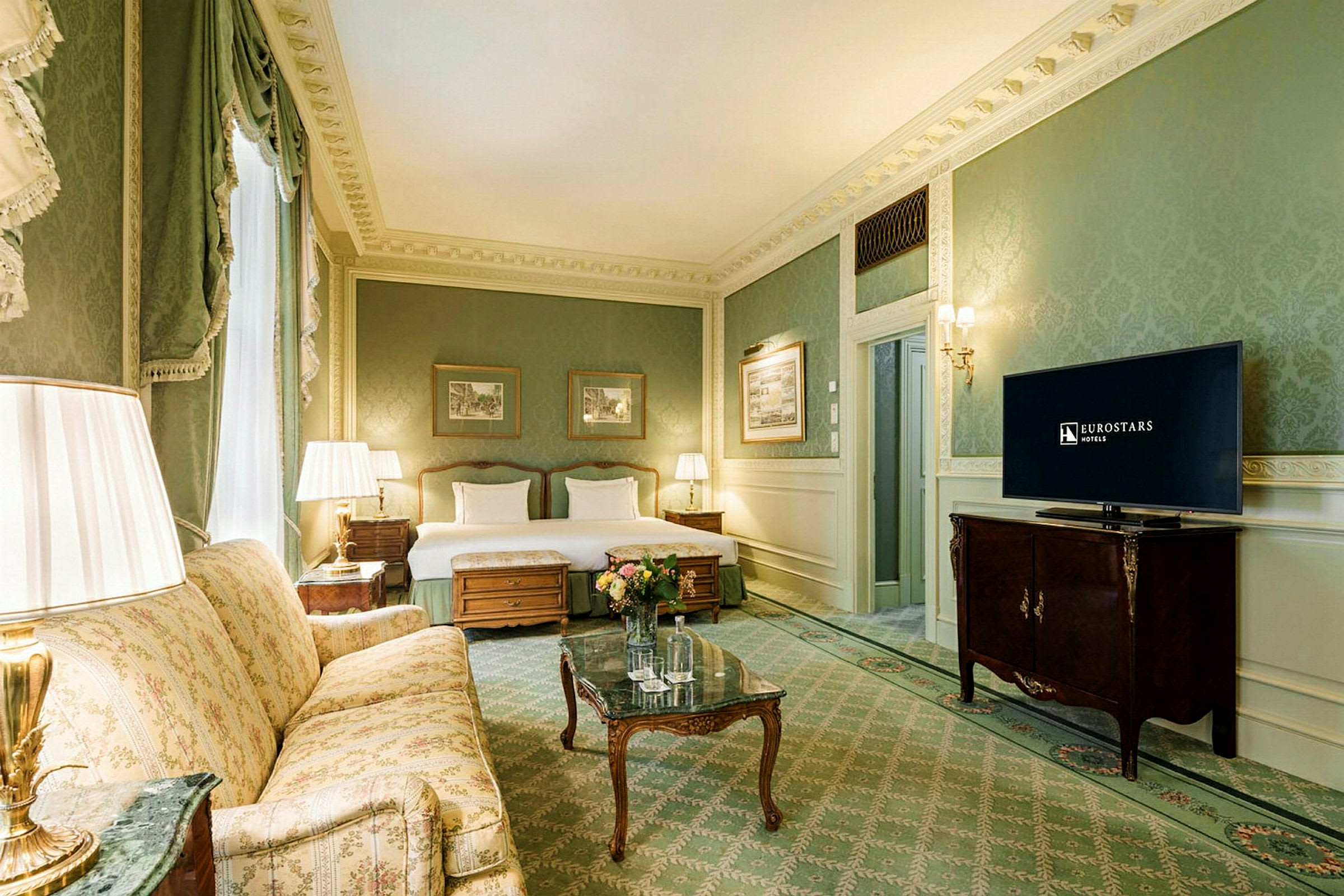 Et hotellrom med klassisk innredning, inkludert en sofa, et bord med blomster, to senger og en TV. (AI-generert tekst)