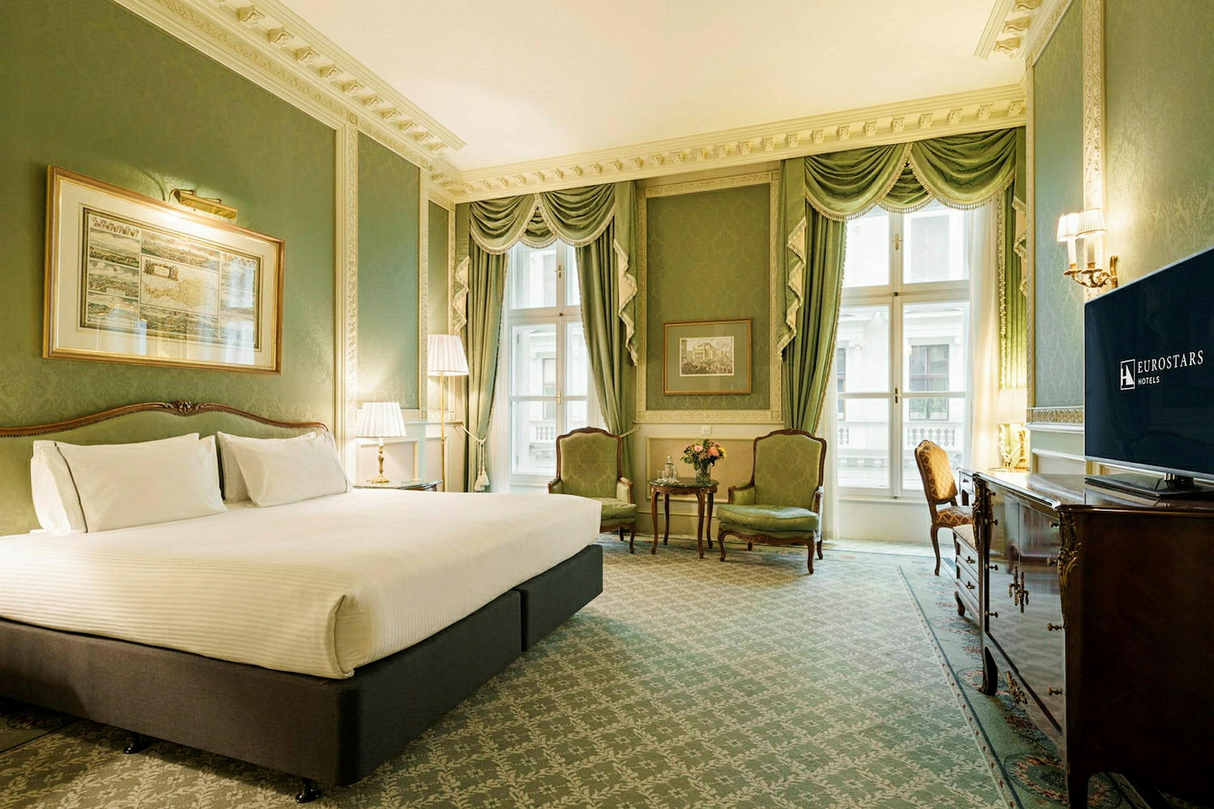Et elegant hotellrom med grønt interiør, en stor seng med hvitt sengetøy, to grønne lenestoler ved et bord og en TV. (AI-generert tekst)