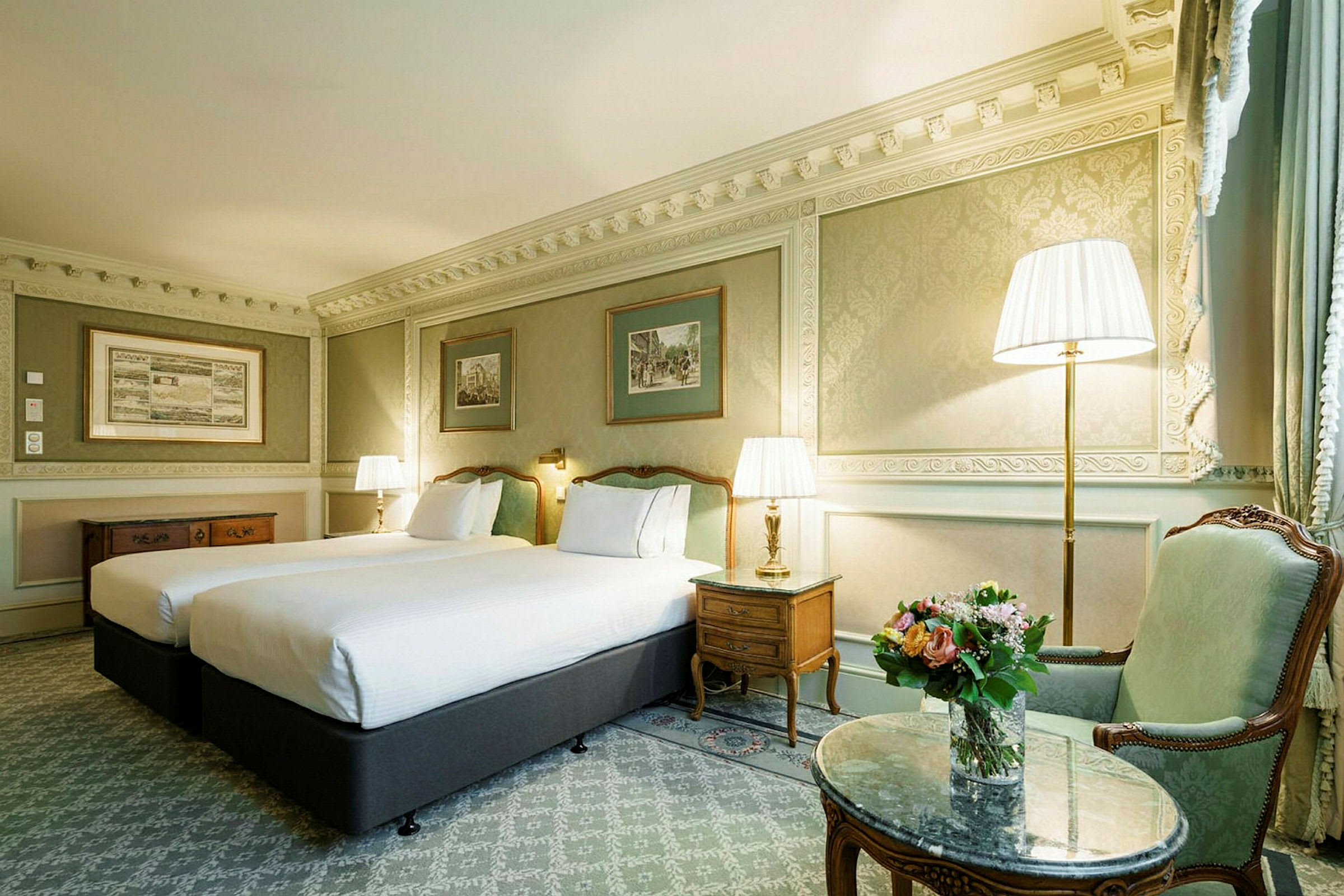 Bildet viser et hotellrom med to enkeltsenger, klassisk innredning og dekorative veggpaneler. Et nattbord med lampe står mellom sengene. En sittegruppe med blomster og marmorbord er synlig til høyre. (AI-generert tekst)