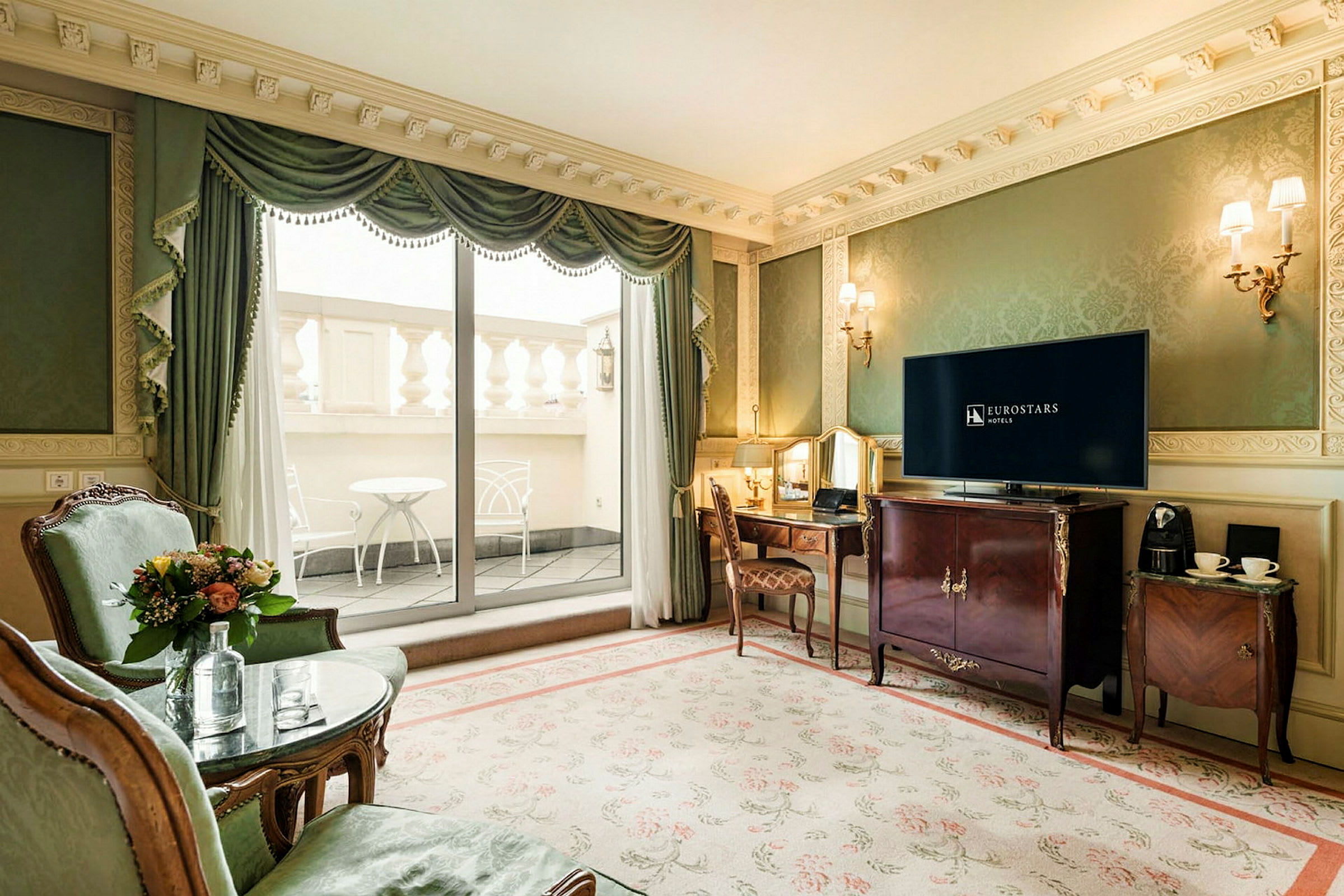 Et elegant hotellrom med grønne vegger, en stor TV, to lenestoler, et skrivebord og en uteplass med møbler. (AI-generert tekst)