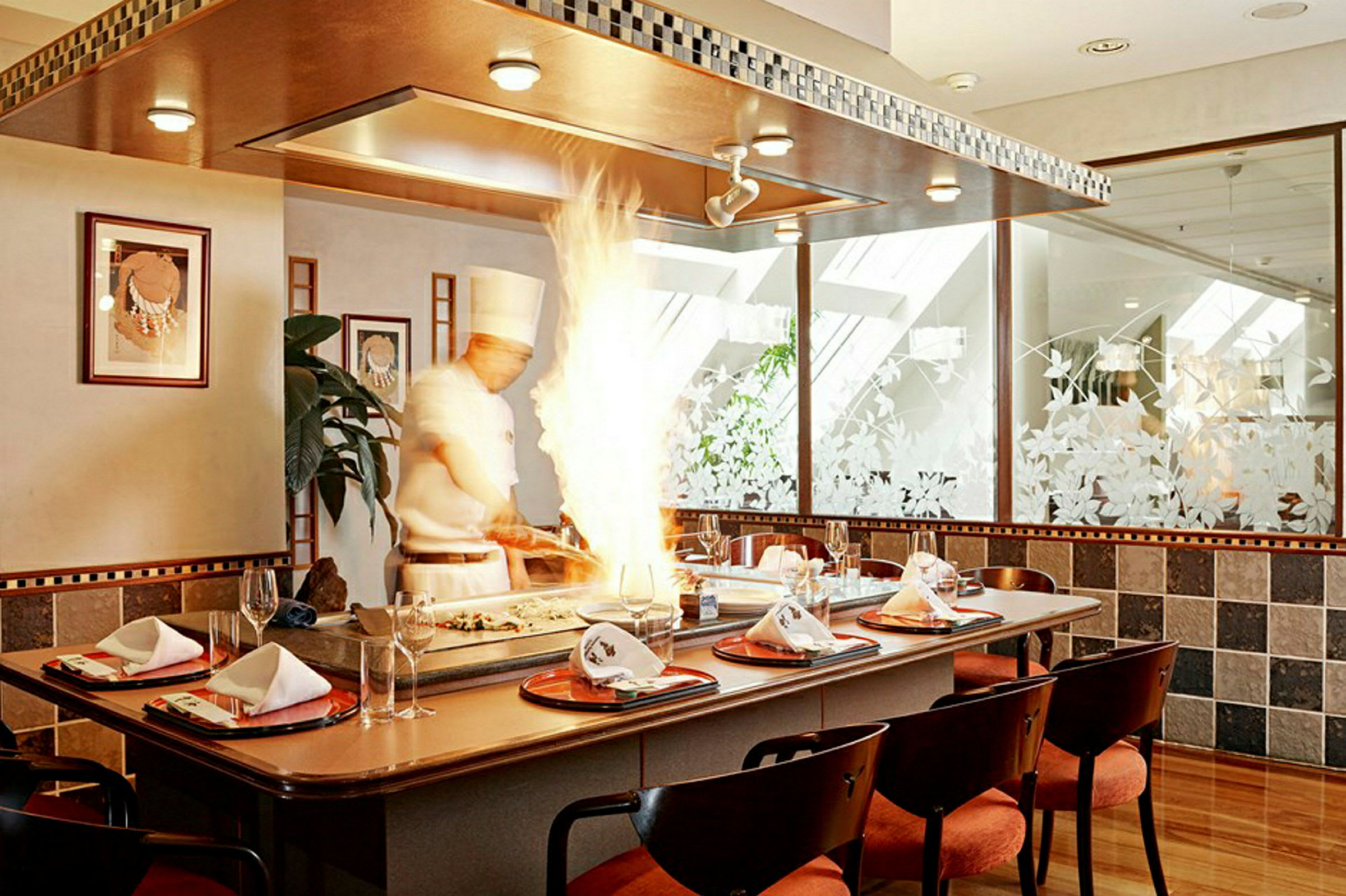 En kokk som arbeider ved en teppanyaki-grill mens flammer stiger opp. Bordet er dekket med servietter og glass. (AI-generert tekst)