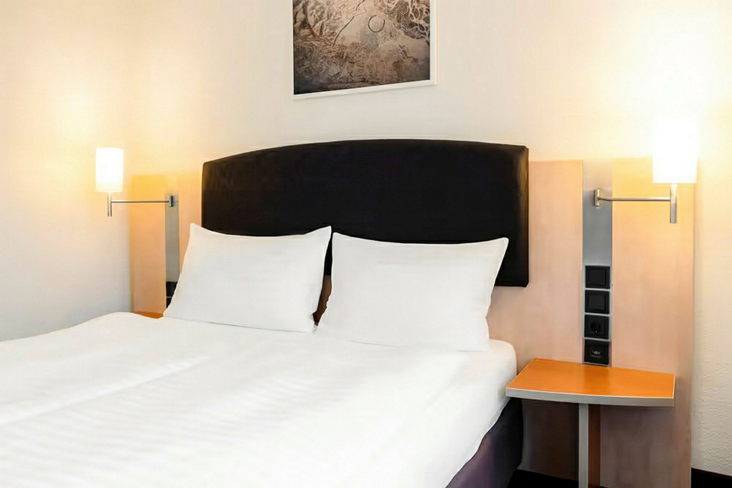 Et hotellrom med en dobbeltseng, hvitt sengetøy og en mørk sengegavl. På begge sider av sengen er det nattbord med lamper festet på veggen. (AI-generert tekst)