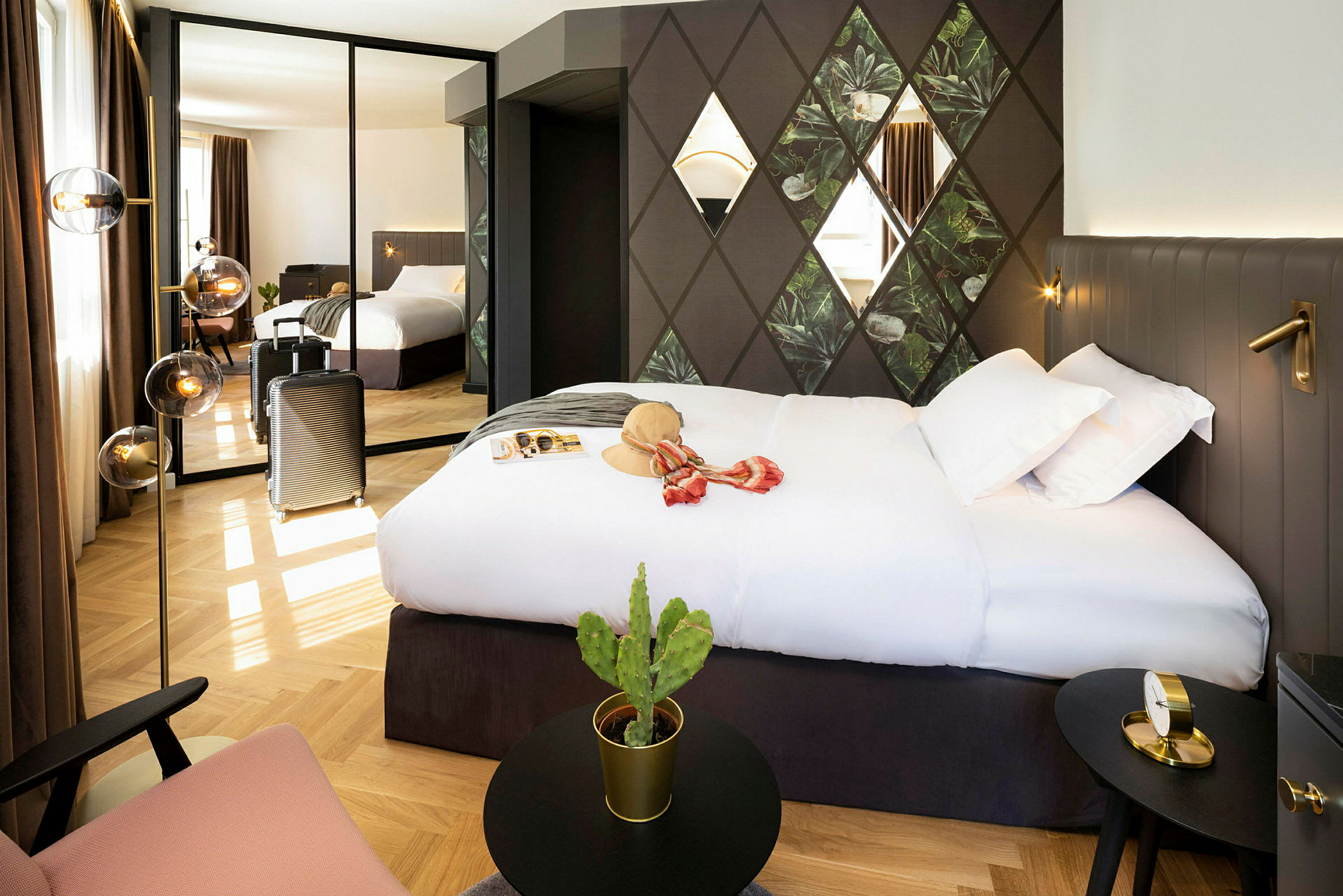 Et hotellrom med en stor seng, veggdekorasjoner og et speil som reflekterer rommet. En koffert står ved siden av, og det finnes en liten kaktus på et bord. (AI-generert tekst)