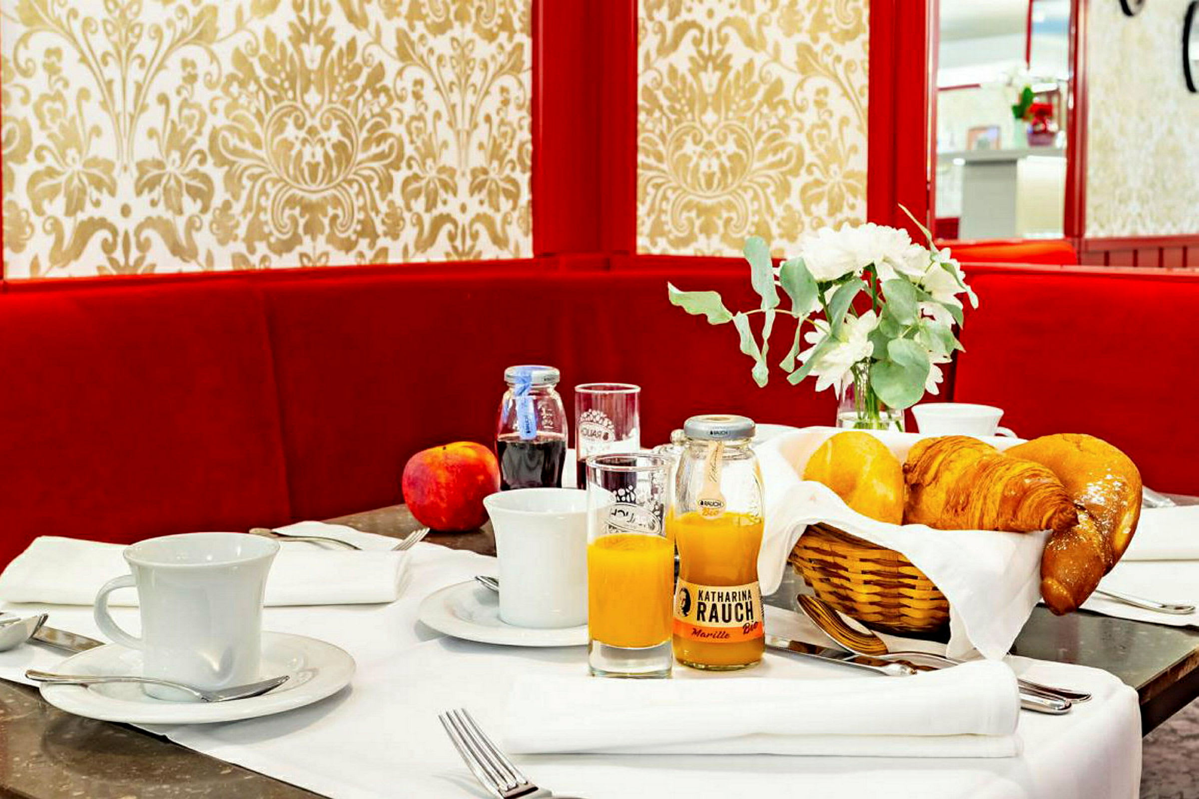 Frokostbord med bakverk, juice, kaffekopper og dekorasjoner mot en rødfarget sofa og mønstret bakgrunn. (AI-generert tekst)