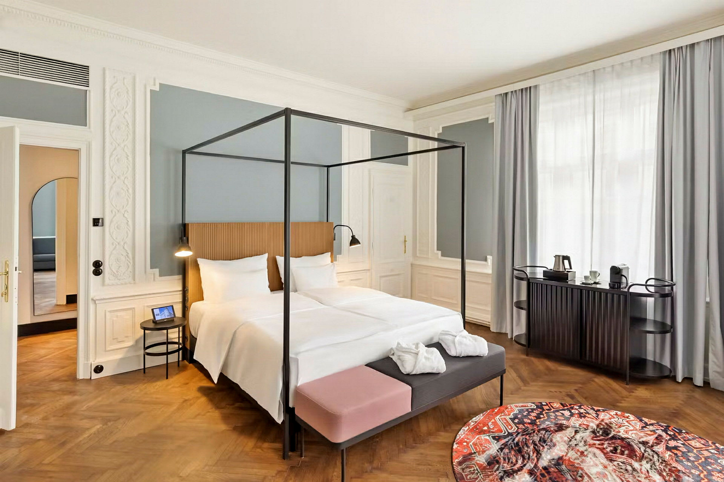 Elegant hotellrom med en himmelseng, et dekorativt rundt teppe og et lite bord samt en kaffestasjon ved vinduet. (AI-generert tekst)