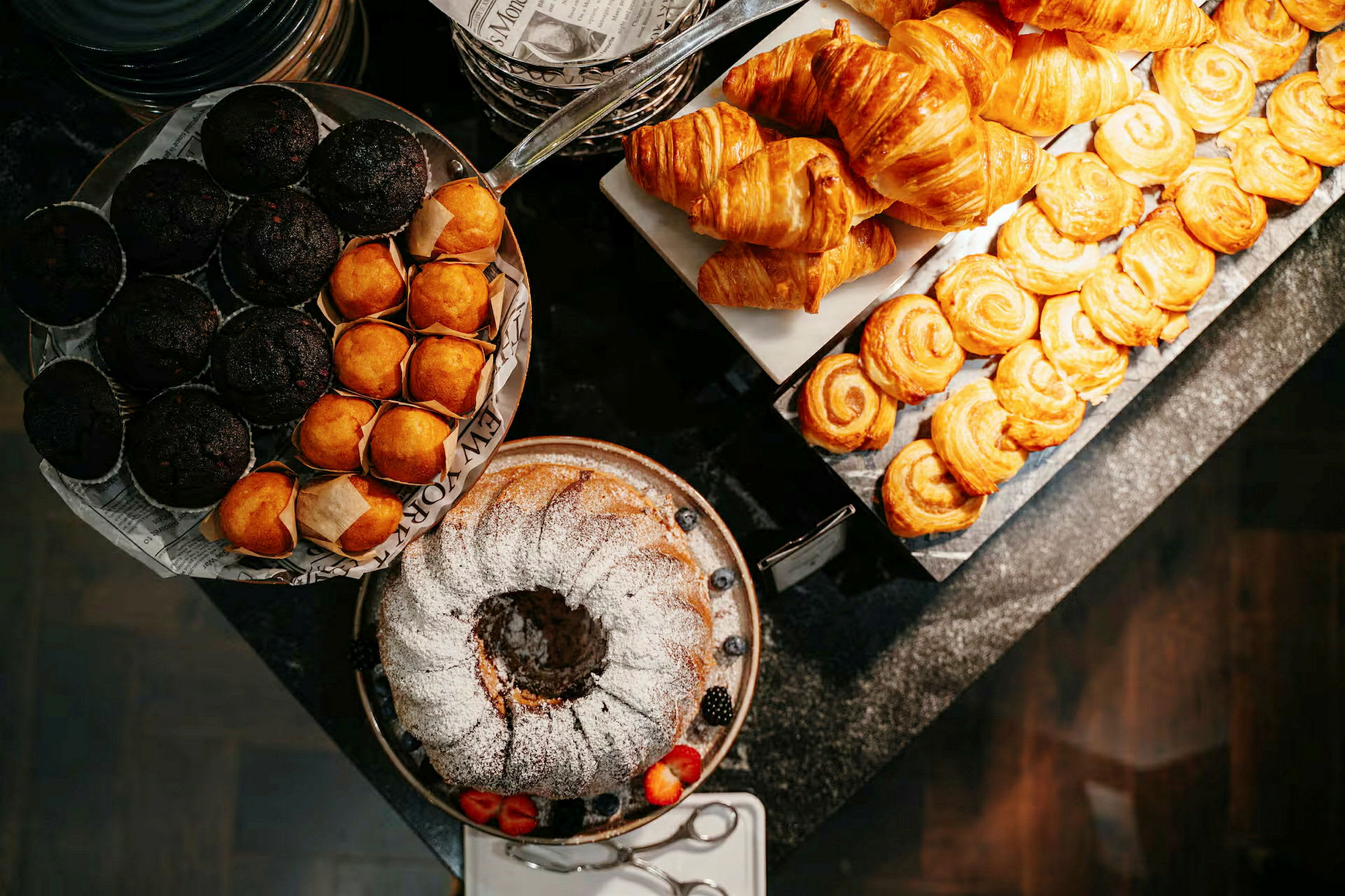 Et utvalg av bakverk som muffins, croissanter og en sukkerkake pyntet med melis, servert på en buffé. (AI-generert tekst)