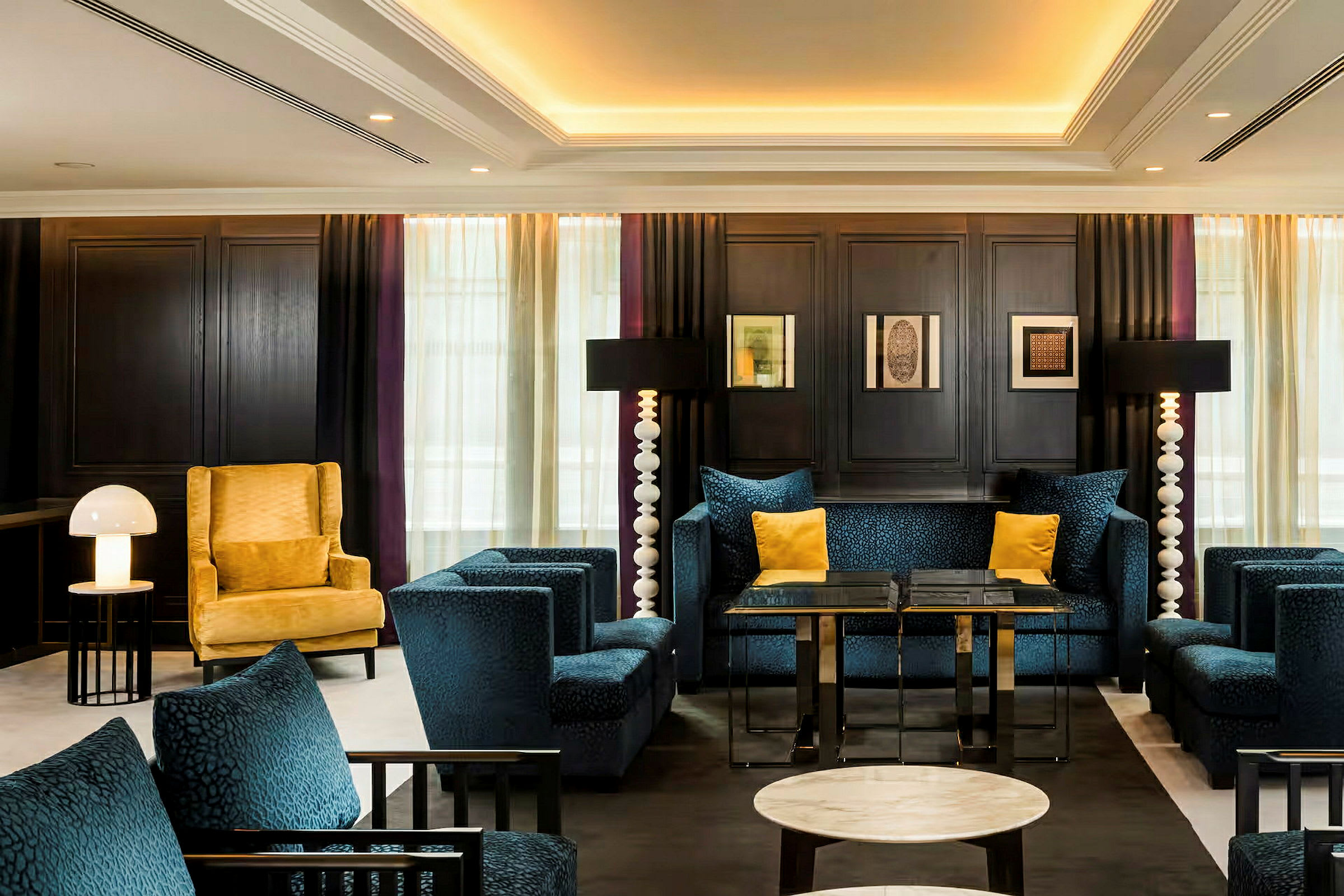 Bilde av et elegant hotellrom eller lounge med blå polstrede møbler, en gul lenestol, bord og lamper. (AI-generert tekst)