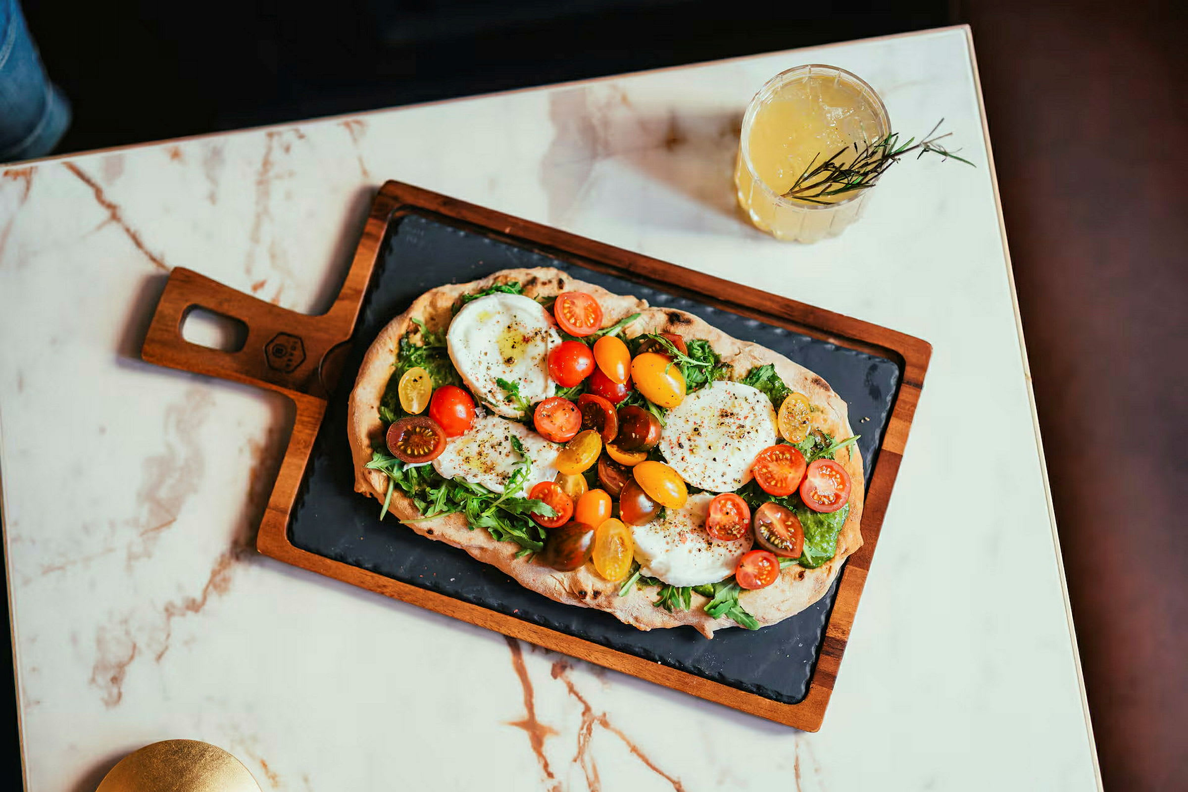 En pizza på et trefat med ruccola, tomater og ost på et marmorbord. En drink står ved siden av. (AI-generert tekst)
