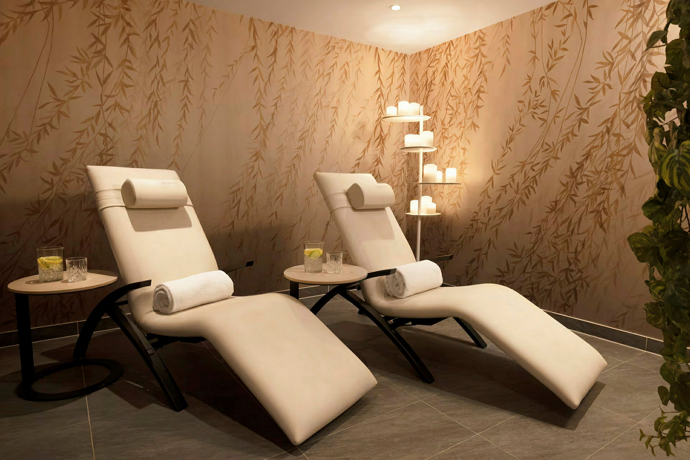 To hvite lenestoler plassert i et avslappende spa-rom med beroligende belysning og en vegg dekorert med plante-motiver. (AI-generert tekst)